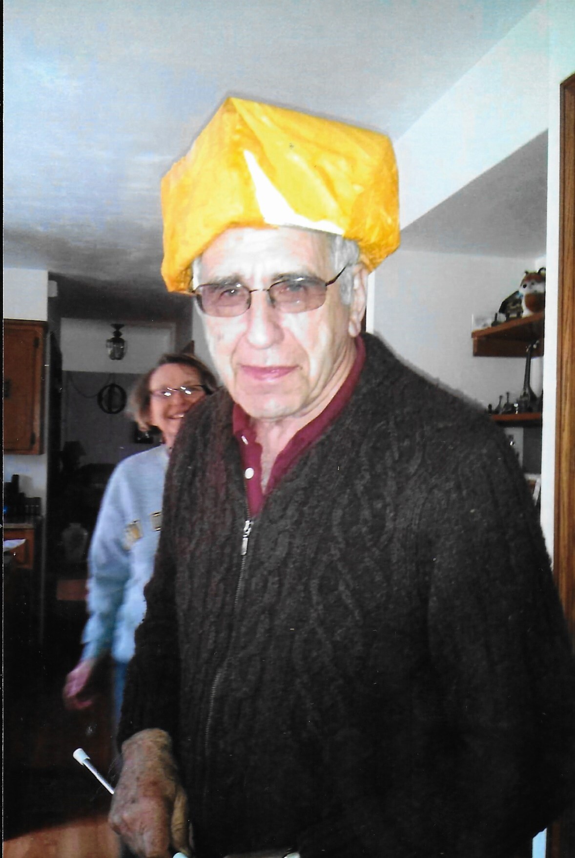 Older man wearing a bright yellow, blocky hat indoors, with a smiling woman in a blue sweater in the background.