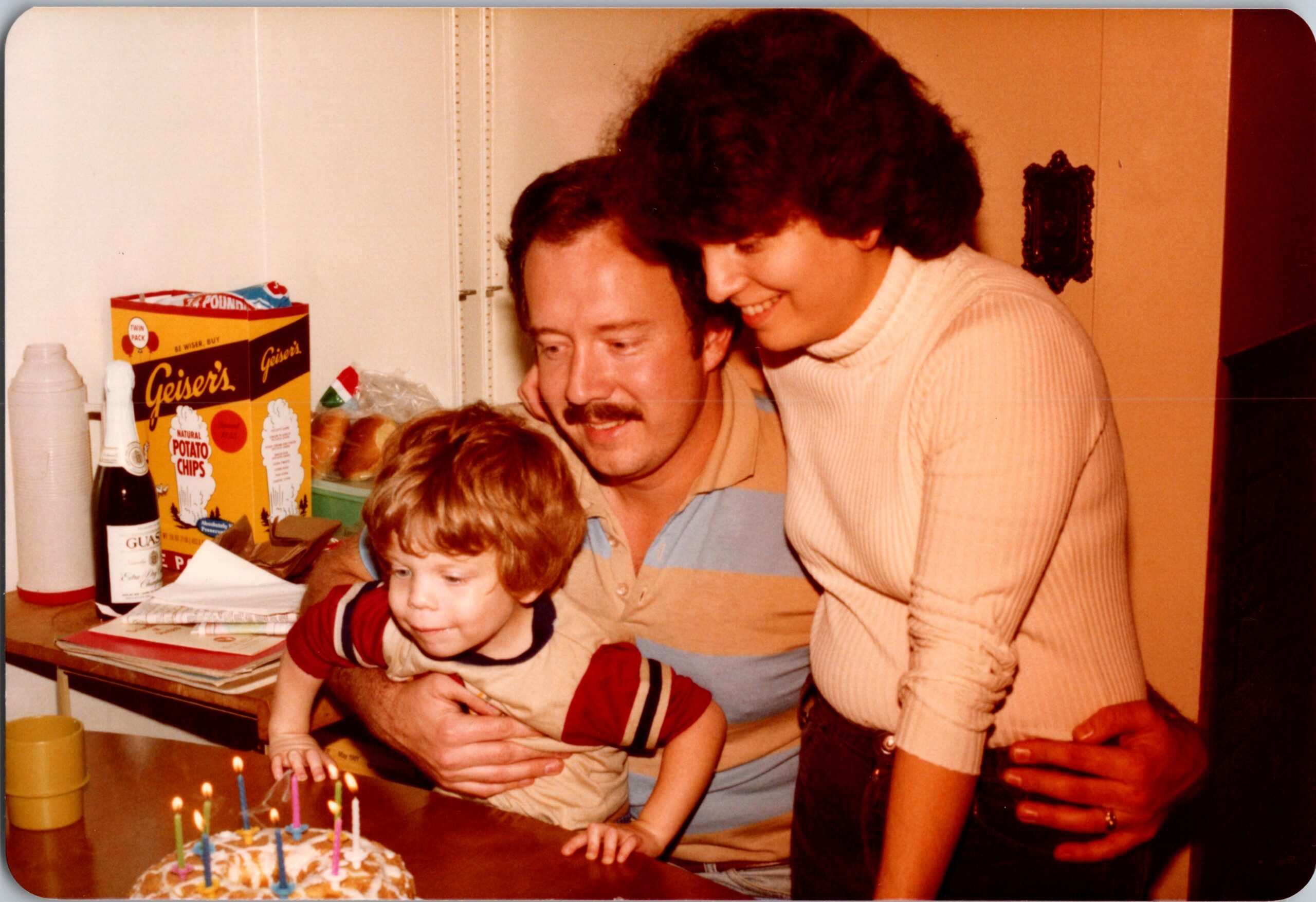 A child leaning over a birthday cake with candles while adults cheer nearby in a kitchen setting.