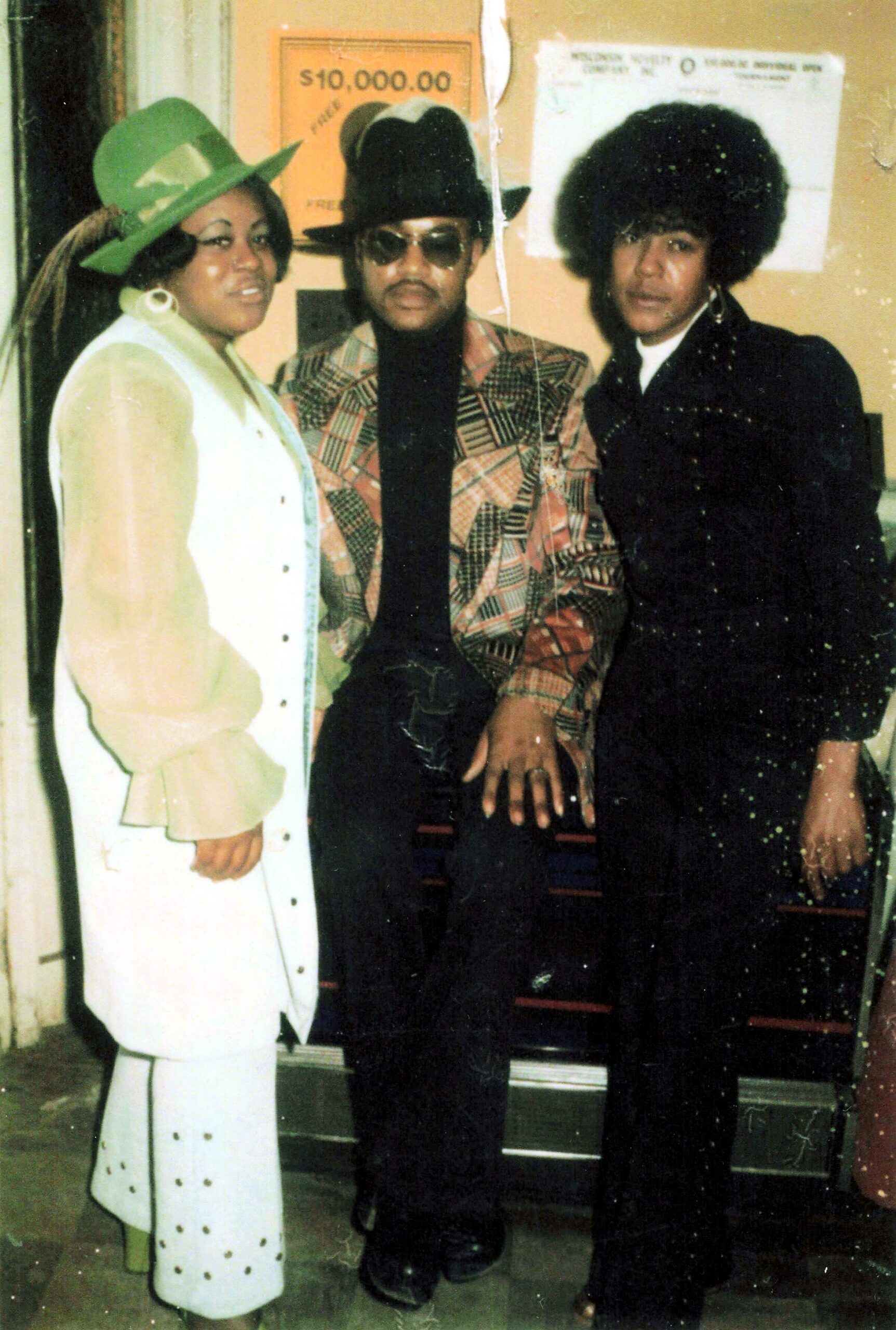 Three people posing in retro outfits, left woman in lime hat and white coat, middle man in plaid blazer and large hat, right person with big afro in dark suit.