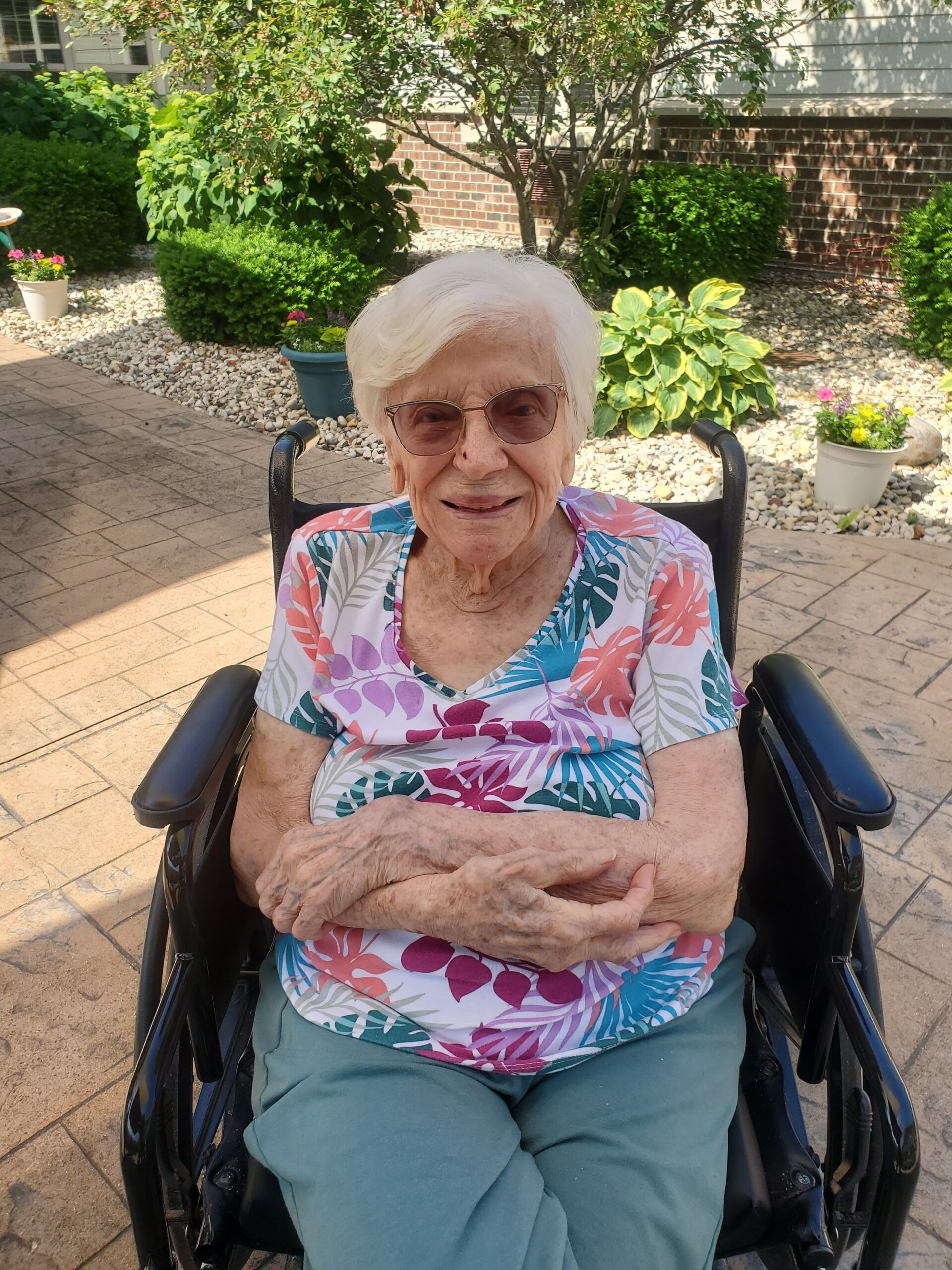 Smiling elderly woman in a wheelchair outdoors in a sunny garden setting with plants behind her.