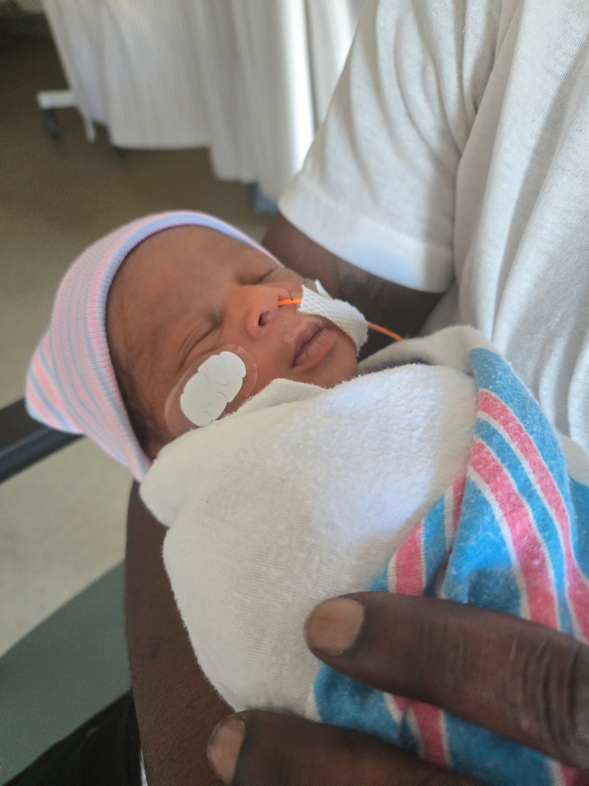 Newborn resting in arms, wrapped in a white blanket with a pastel striped hat and nasal tubing taped to the cheek.
