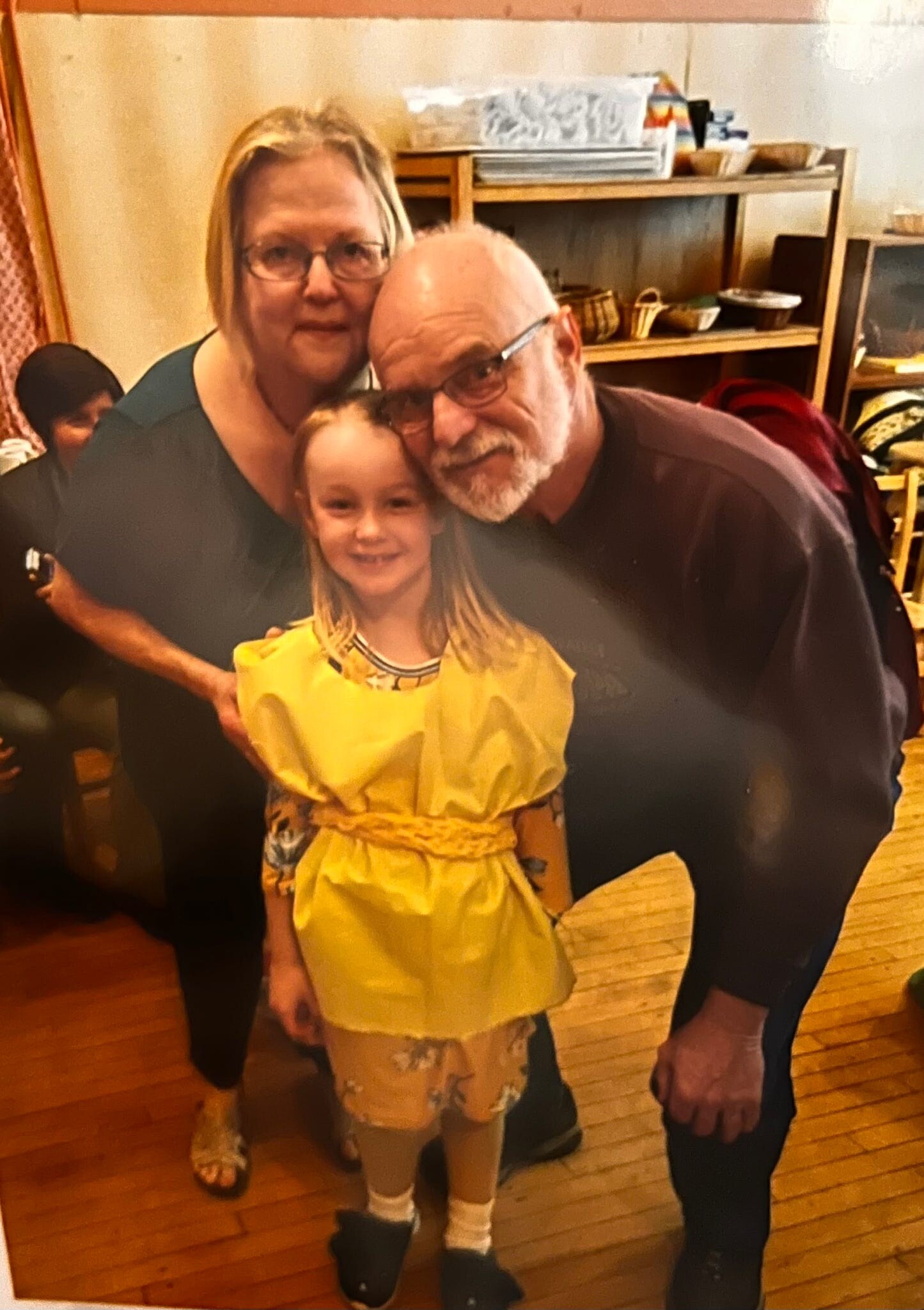 Family group indoors: grandfather with glasses leans toward a smiling young girl in a yellow dress, with a woman beside them as they pose for a photo.