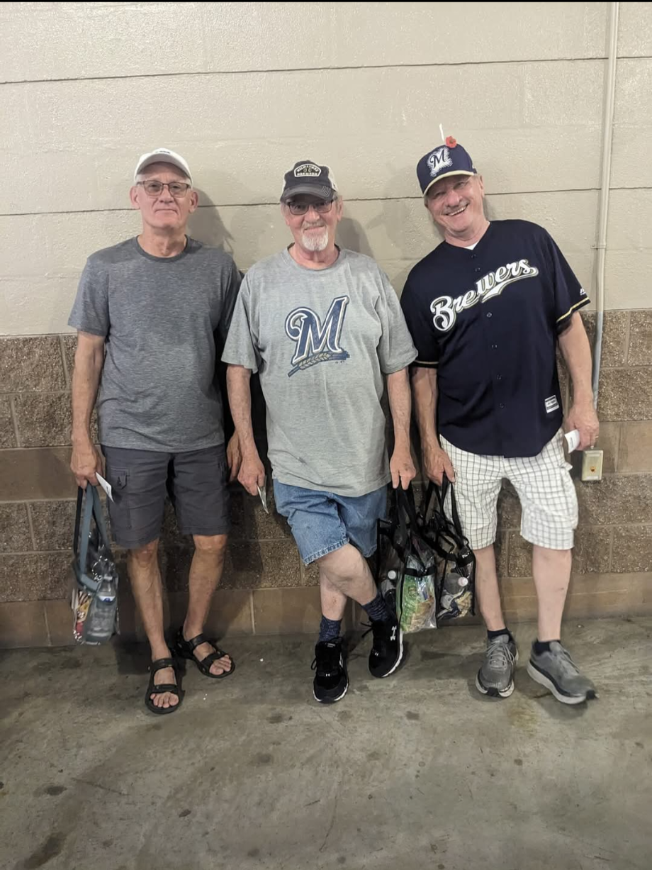 Three older men stand against a beige cinder block wall, each holding a bag of snacks, casually dressed with Brewers gear on two of them.