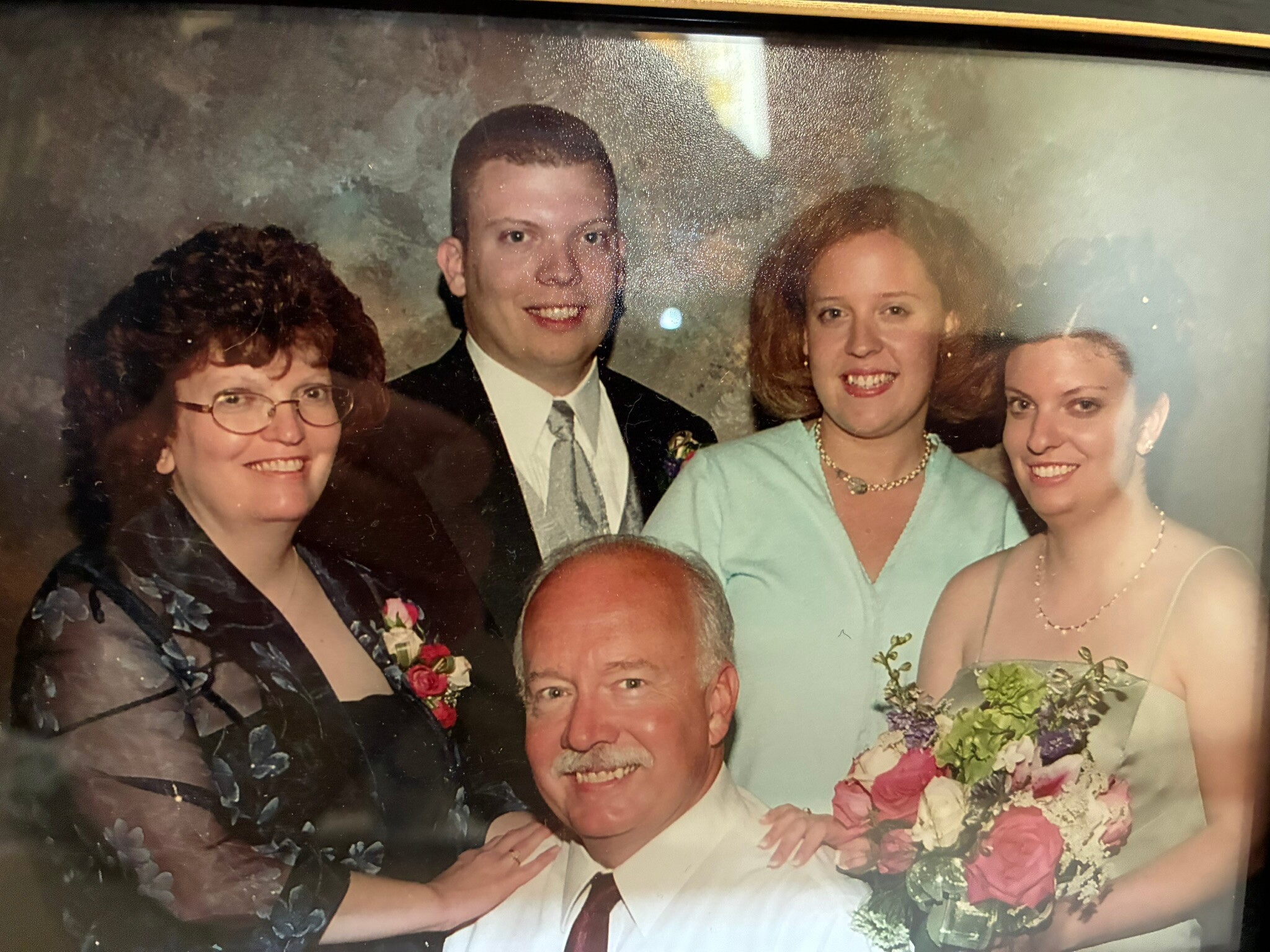 Group wedding portrait: smiling man in a white suit flanked by women in dresses, bouquet visible in foreground at bottom right.