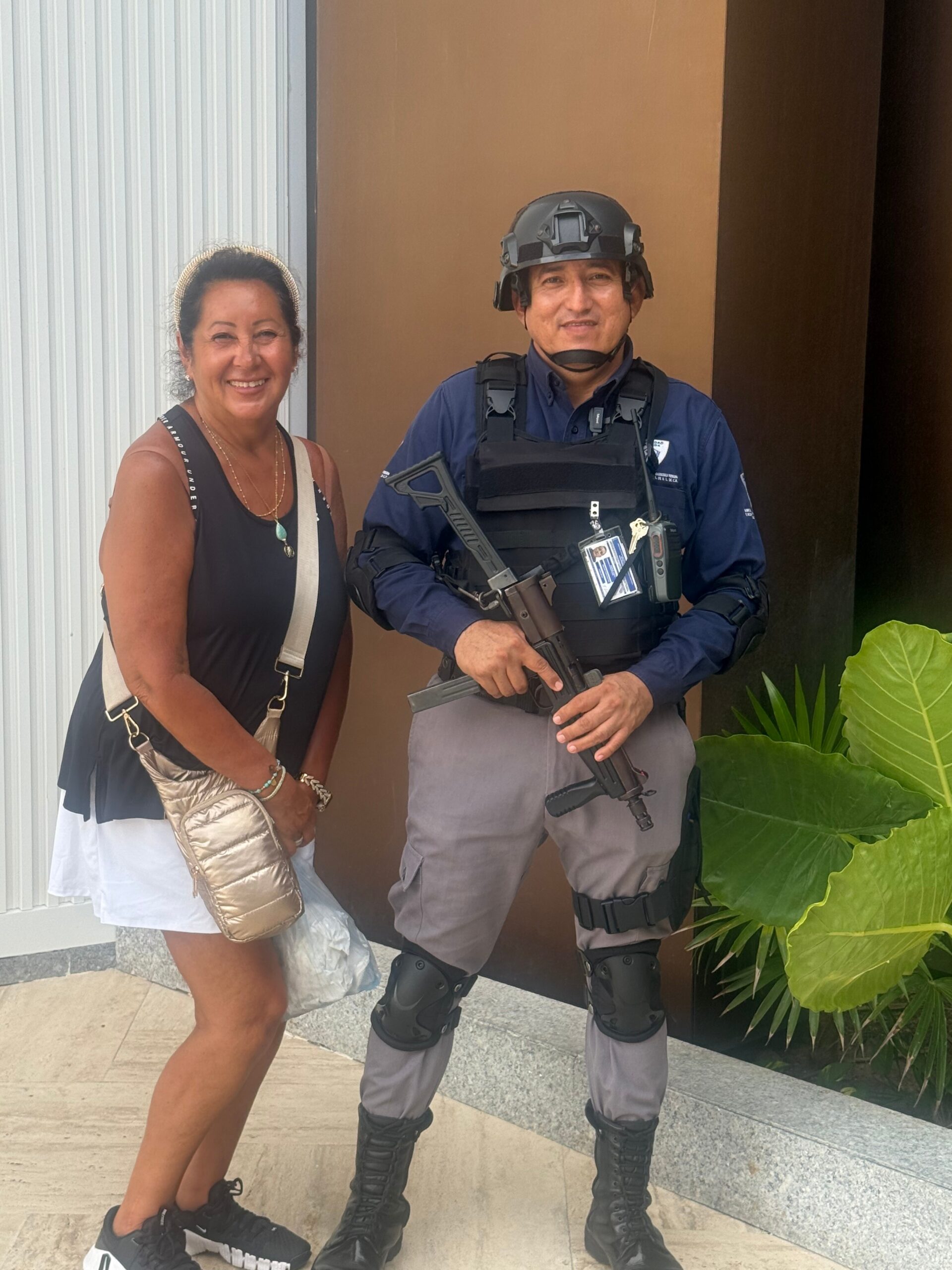 Smiling woman in a black sleeveless top and white skirt standing next to a man in tactical gear with a helmet, body armor, and a rifle.