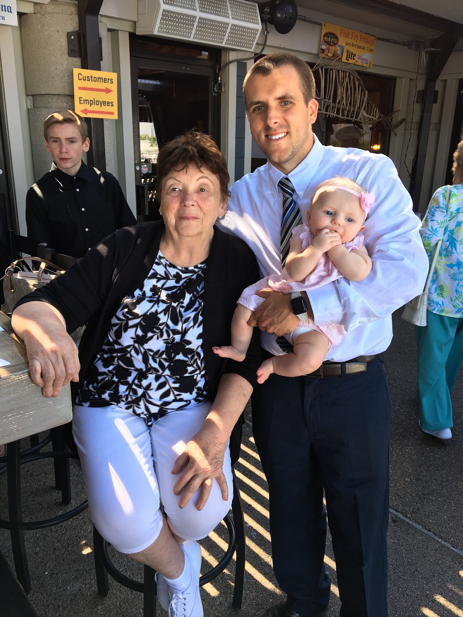 Man in a light blue shirt holds a baby girl in a pink dress while an older woman sits beside them at an outdoor restaurant.