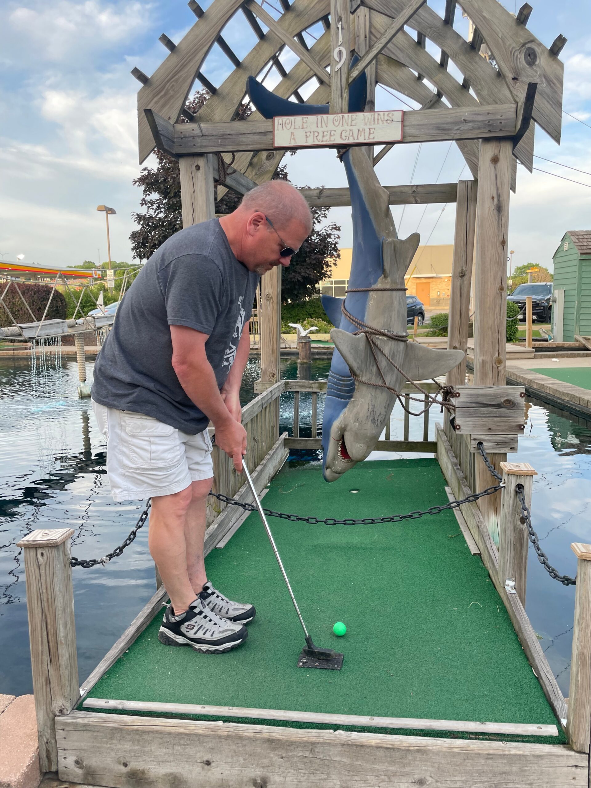 Man lining up a putt on a mini-golf green with a shark sculpture and wooden frame behind him; ball nearby and a sign reading 'Hole in one wins a free game'