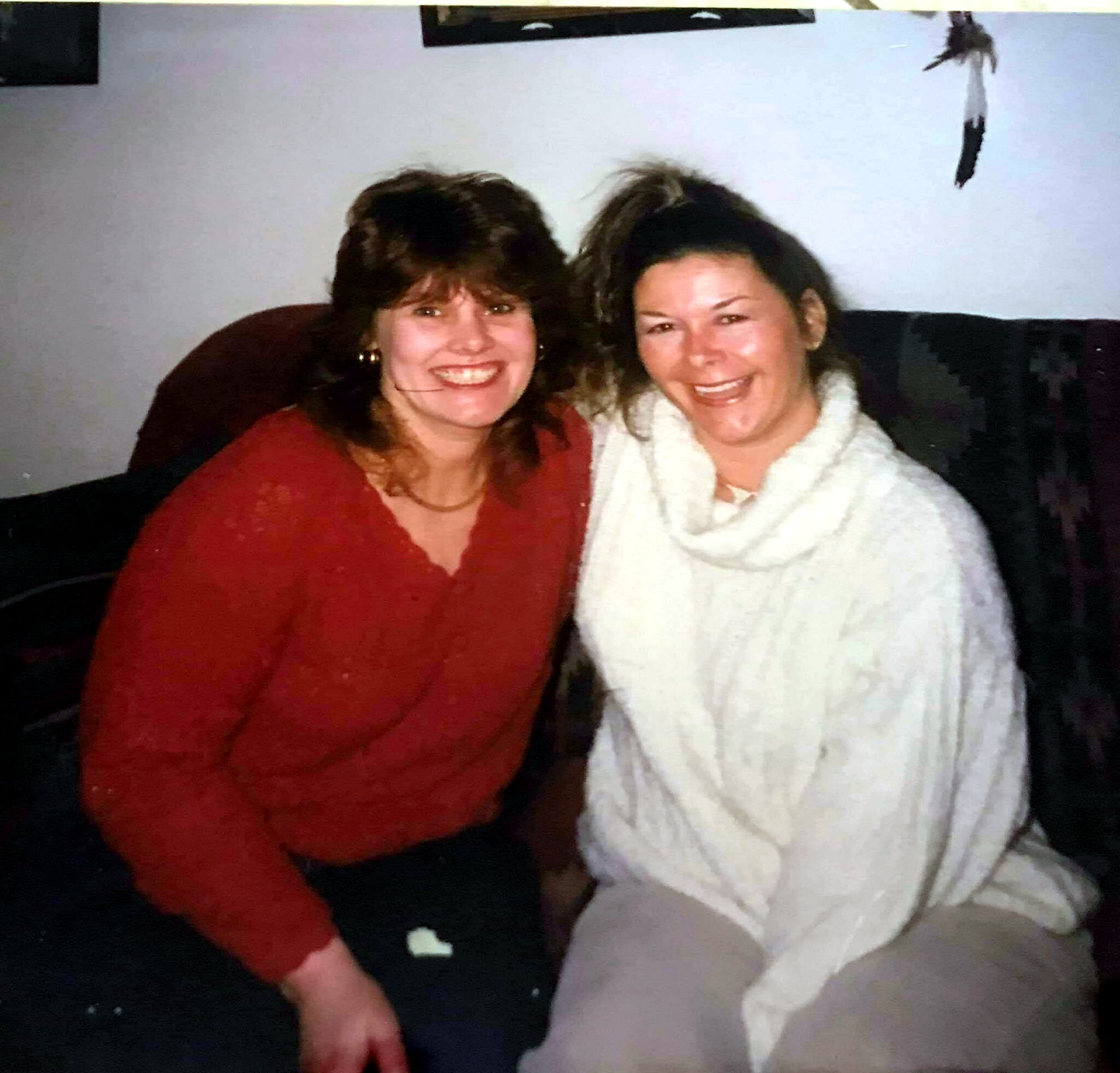Two women sit close together on a couch, smiling at the camera, one in red and the other in white.
