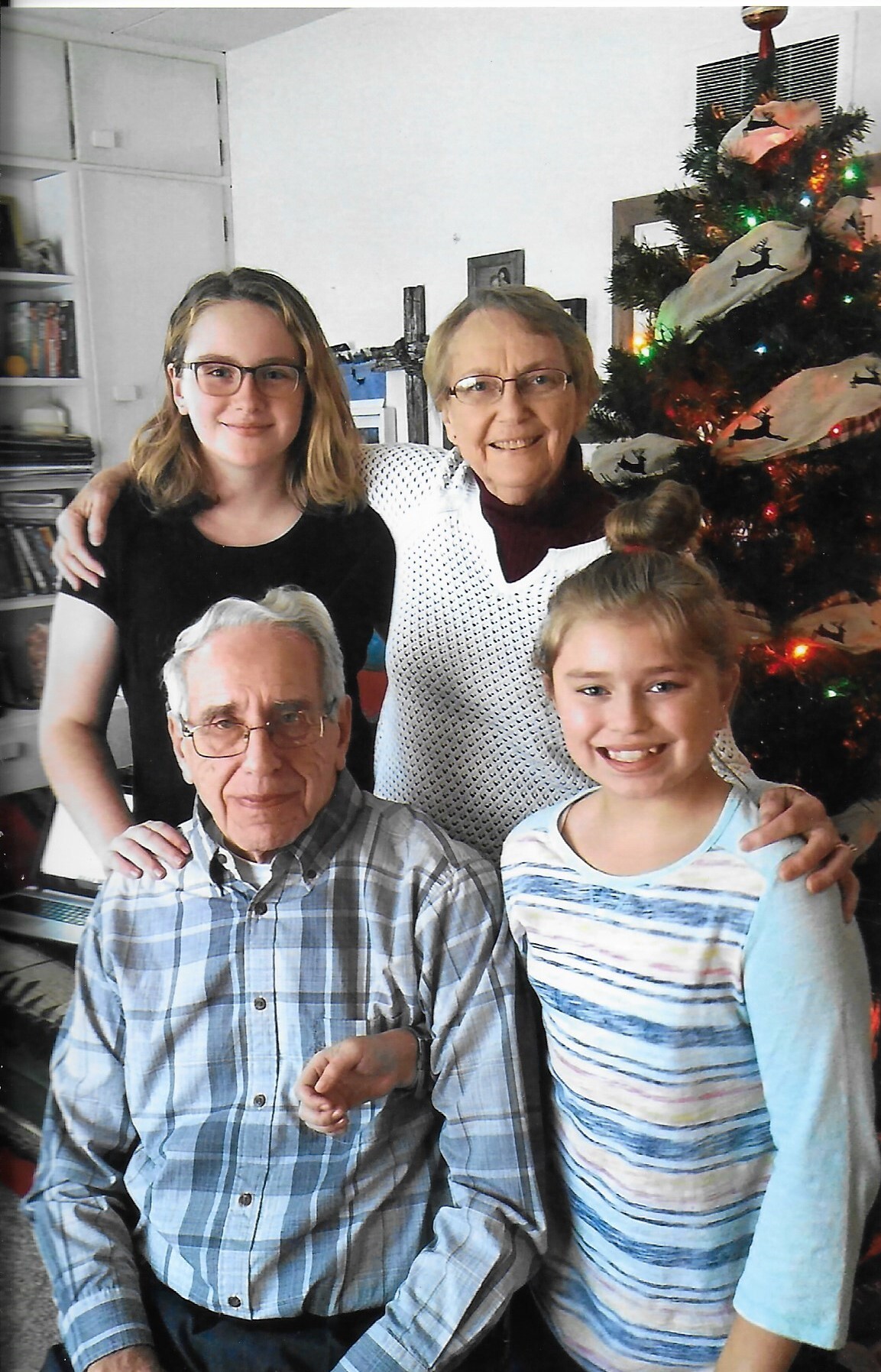 Family group portrait in a living room with a Christmas tree: an elderly man seated, two girls, and a smiling woman standing behind them.