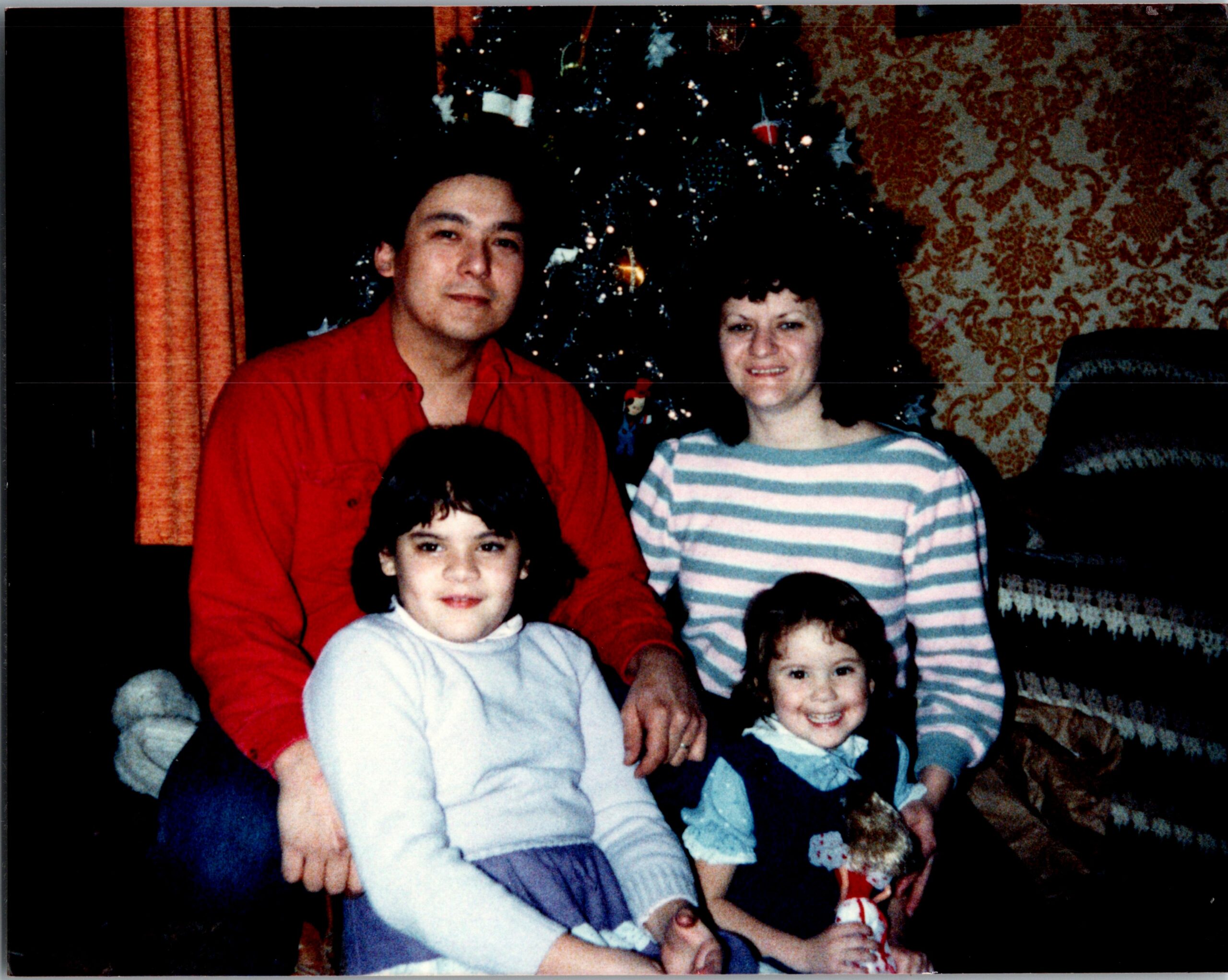 Family group posing for a holiday photo in front of a Christmas tree, smiling faces and festive sweaters/overtop outfits visible.