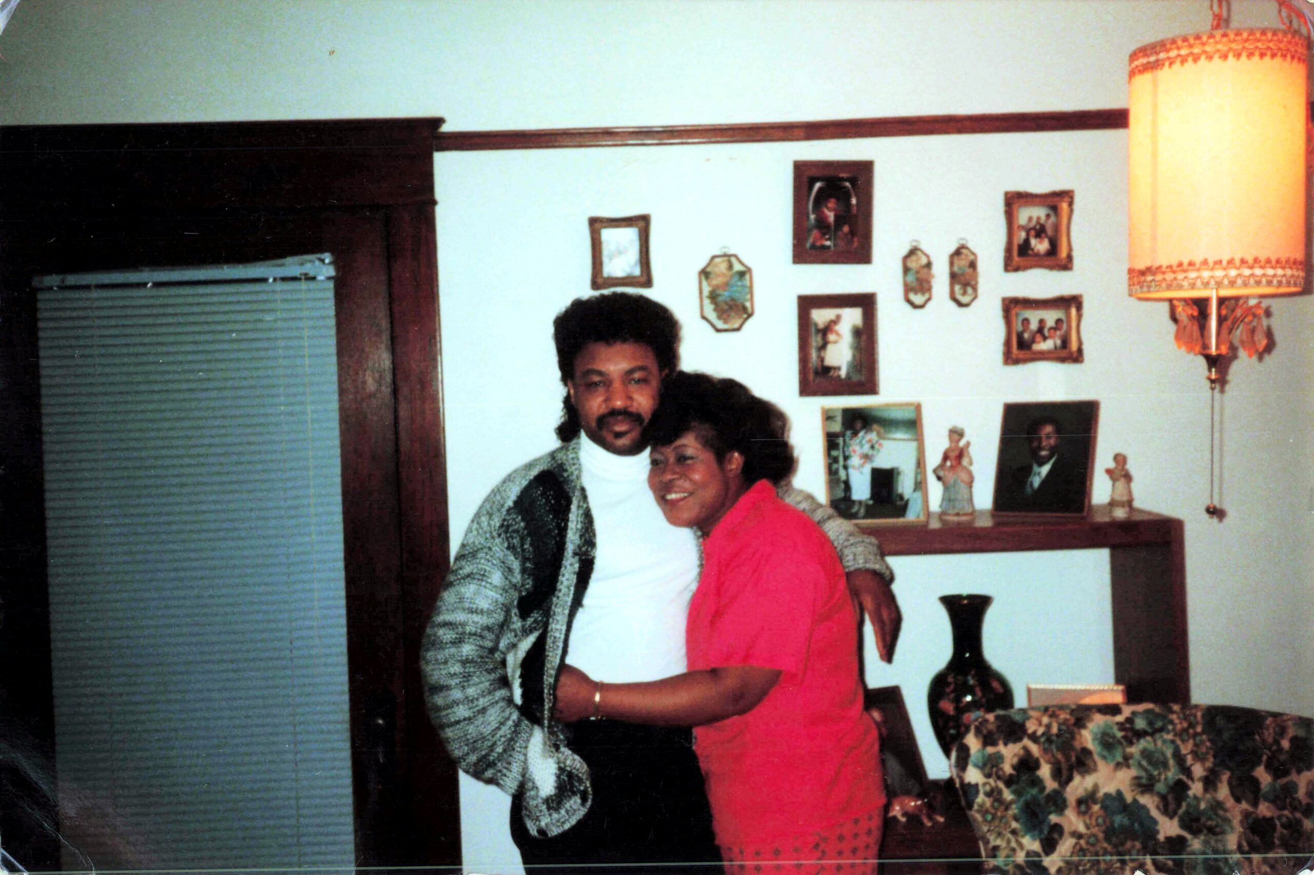 Couple smiling and hugging in a living room with a wall full of family photos and a tall lampshade nearby.