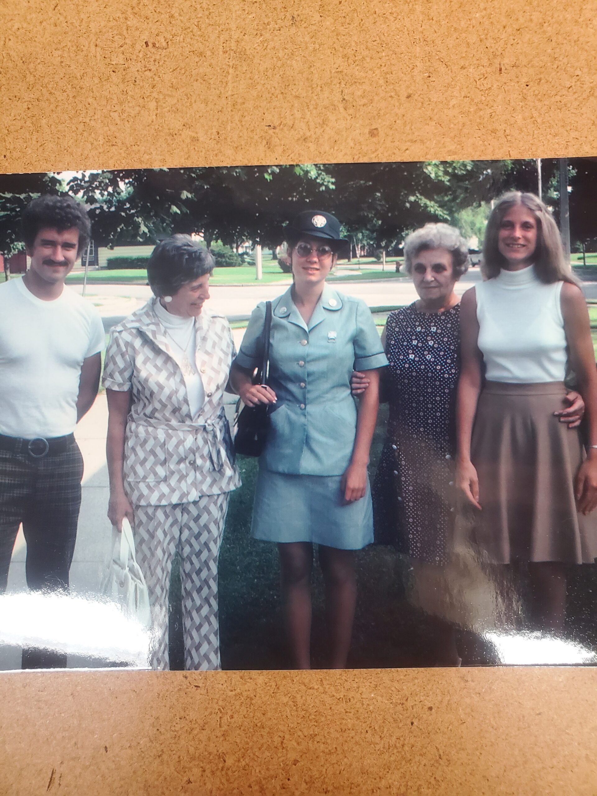 Five people stand together outdoors on a sunny day, posing for a photo with trees and a lawn in the background.