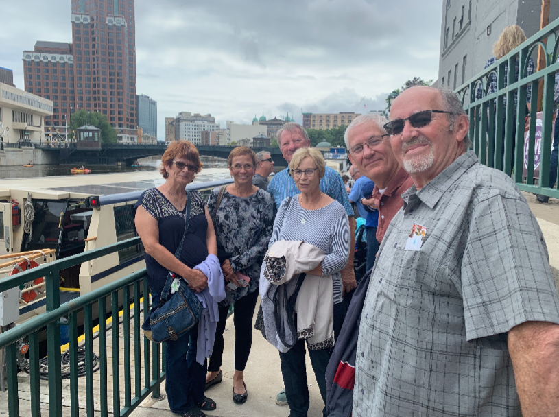 Group of smiling seniors posing on a riverside promenade with a docked boat and city buildings in the background, casually dressed for a daytime outing.