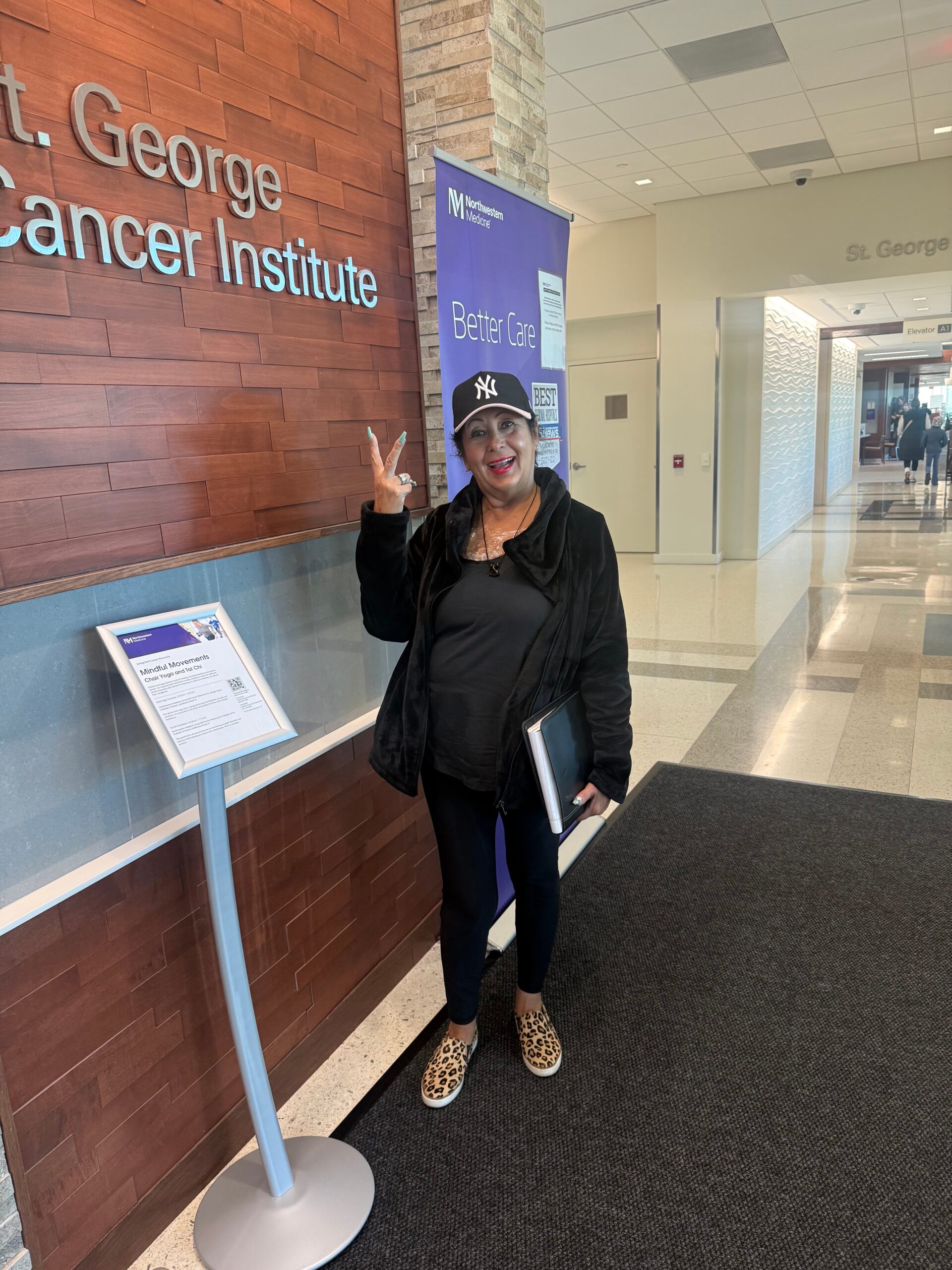 Woman in black jacket and leopard shoes poses with peace sign in hospital lobby beside St. George Cancer Institute sign and a blue Better Care banner.