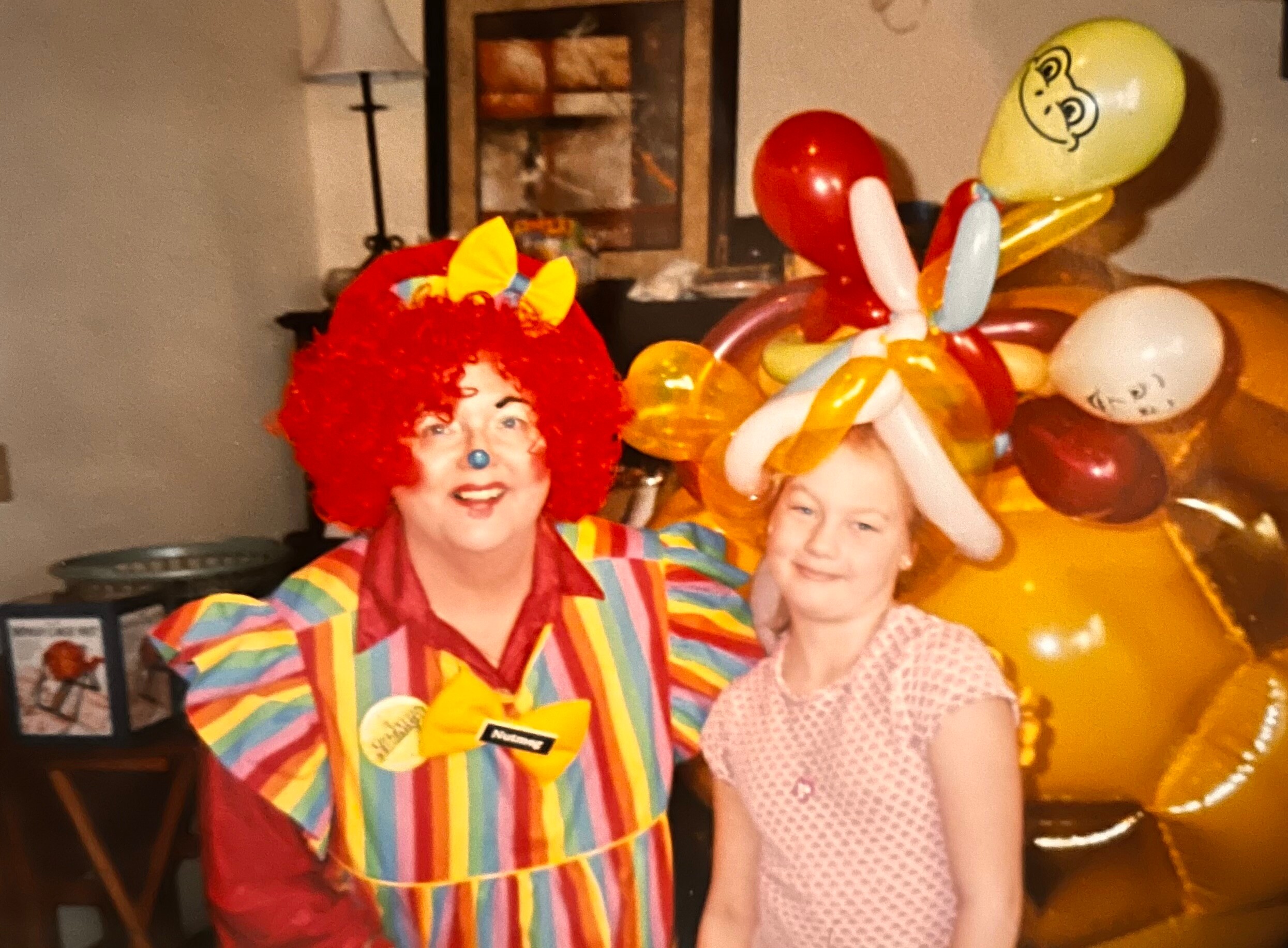 Two people in clown costumes posing indoors with colorful balloon decorations in the background stride a festive party scene