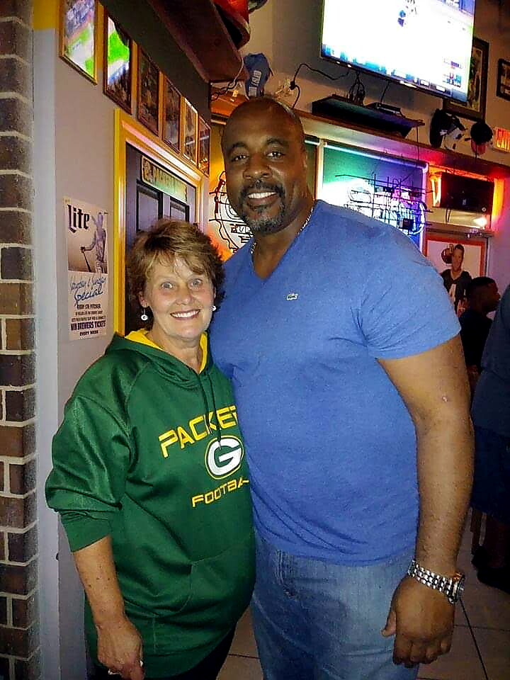 Smiling woman in a green Packers hoodie posing with a tall man in a blue t-shirt at a neon-lit bar.