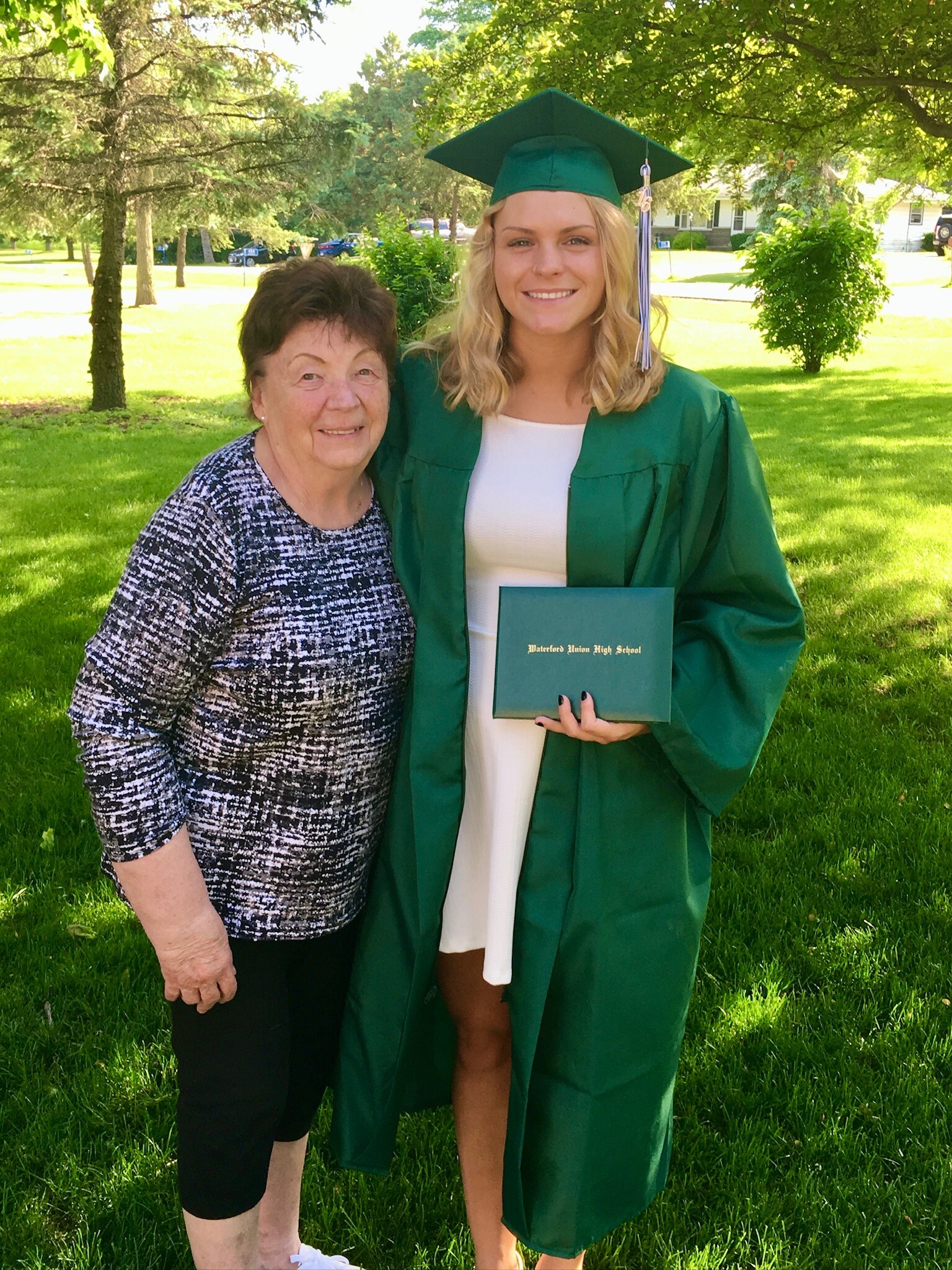 Graduate in green cap and gown posing with an older woman outdoors, holding a green diploma cover.