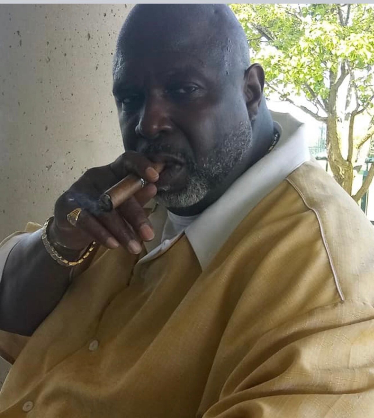 Man smoking a cigar outdoors, wearing a tan shirt and gold jewelry (watch, rings) with a gray beard.