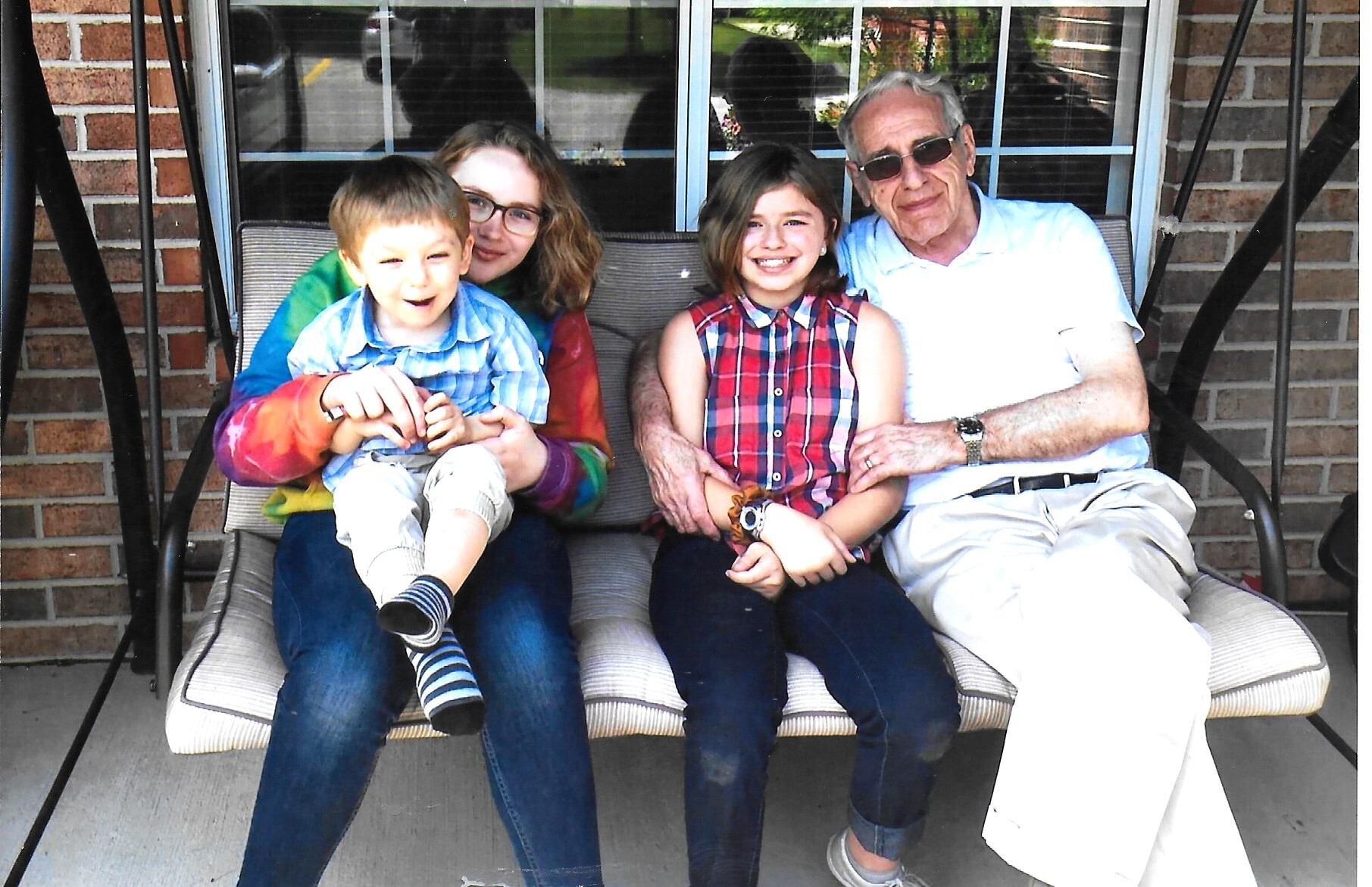 Grandfather in sunglasses sits with grandmother and two kids on a porch swing, smiling for a family photo.