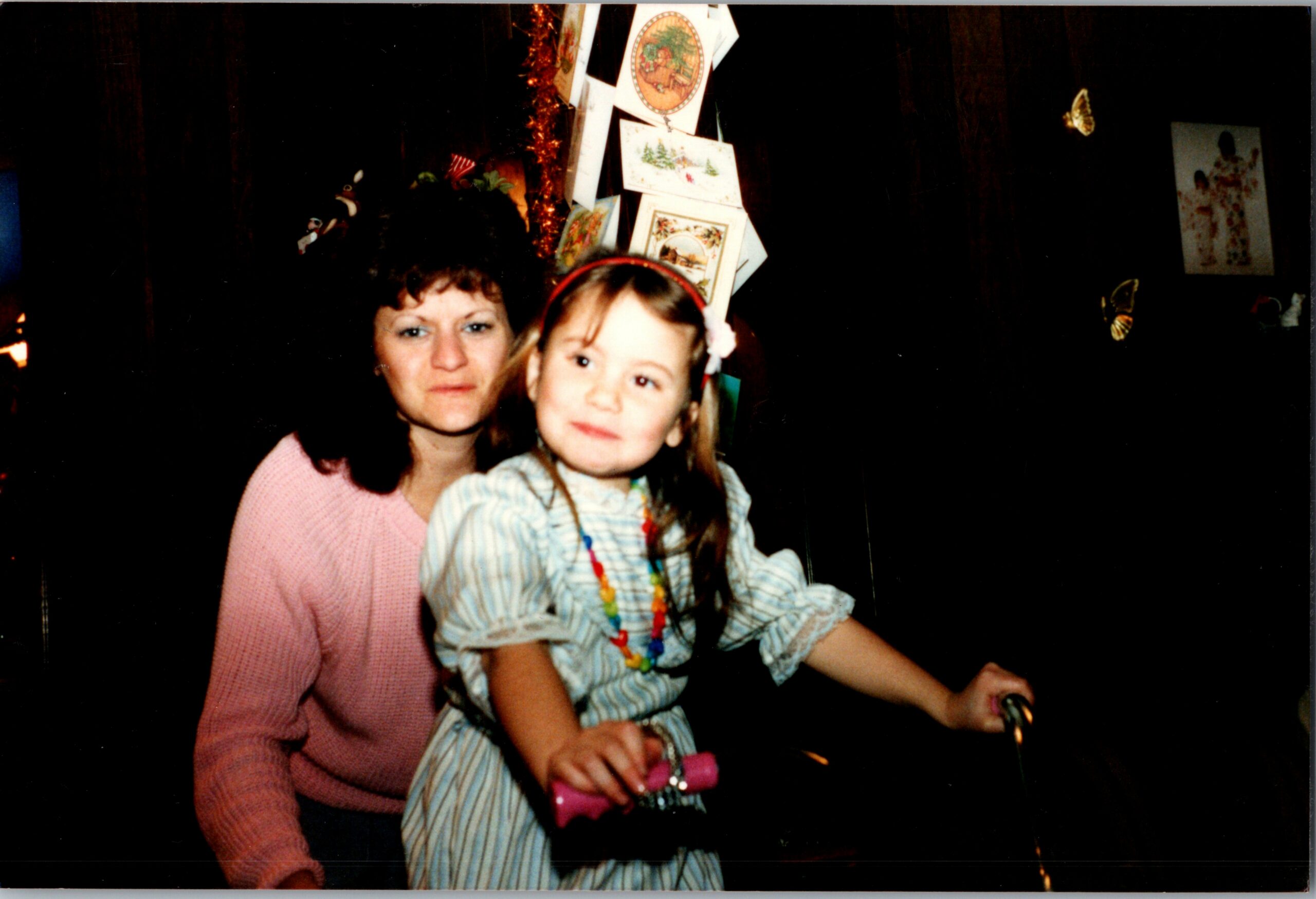 Woman and young girl posing together at a holiday party, Christmas cards hanging overhead in a dark room.