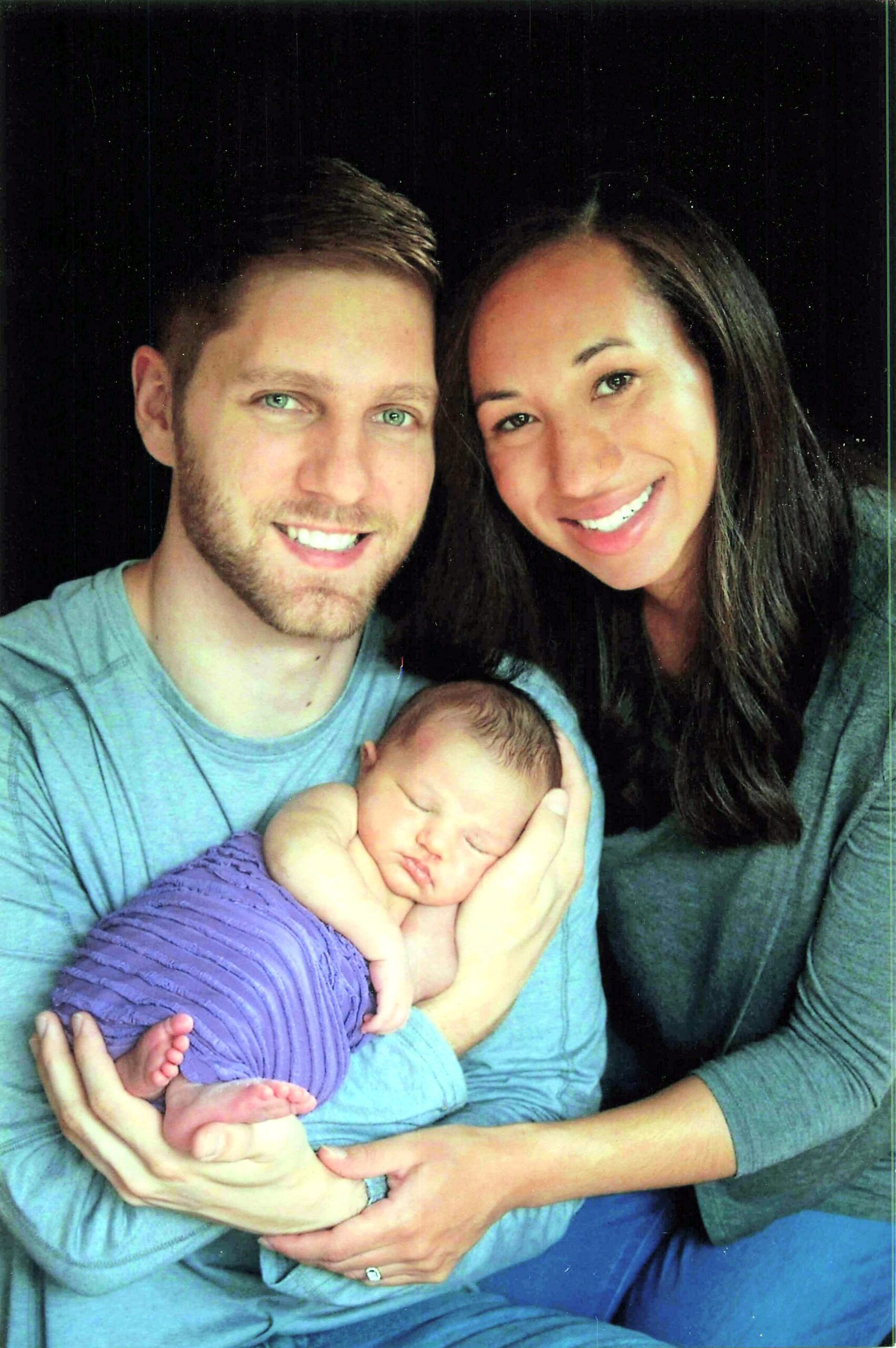 Couple with their newborn baby cradled in the parents' arms, all smiling at the camera.