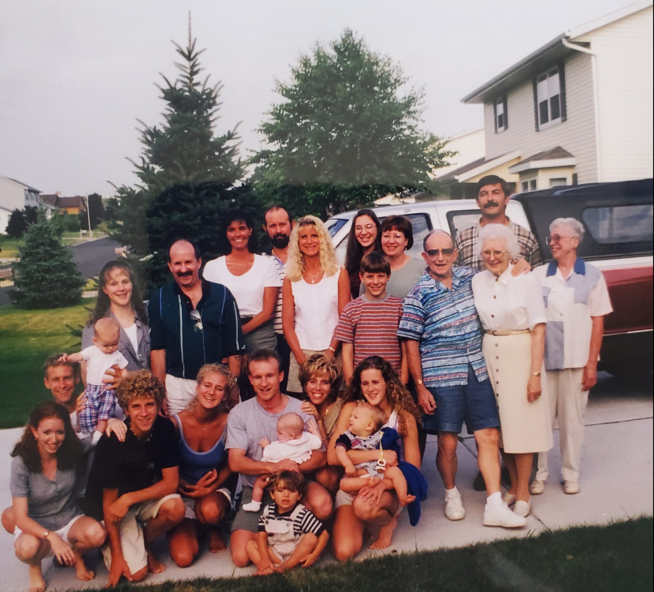 Large, multi-generational family group posing outdoors in front of a white truck in a suburban yard, with a two-story house and trees in the background.