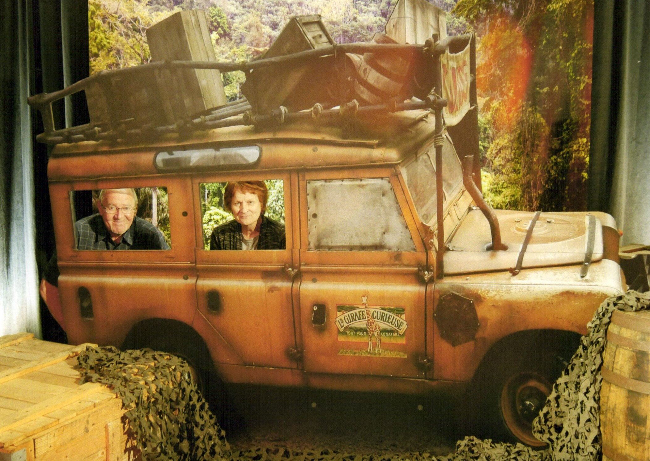 Orange vintage bus on display with two people sitting in the front seats, surrounded by camouflage netting and a forest backdrop.