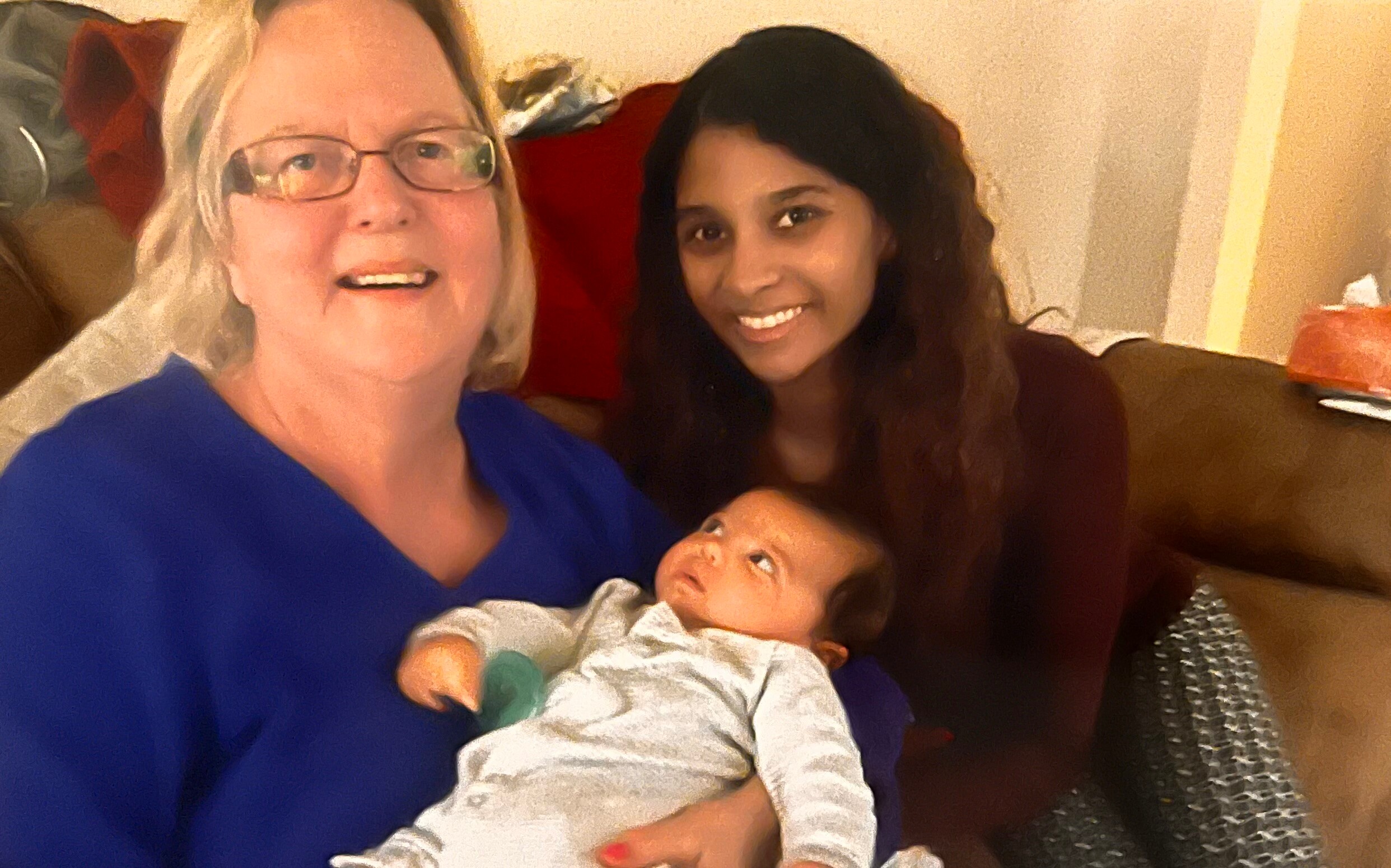 Grandmother wearing blue holds a baby while a young woman sits beside on a sofa, all smiling at the camera.