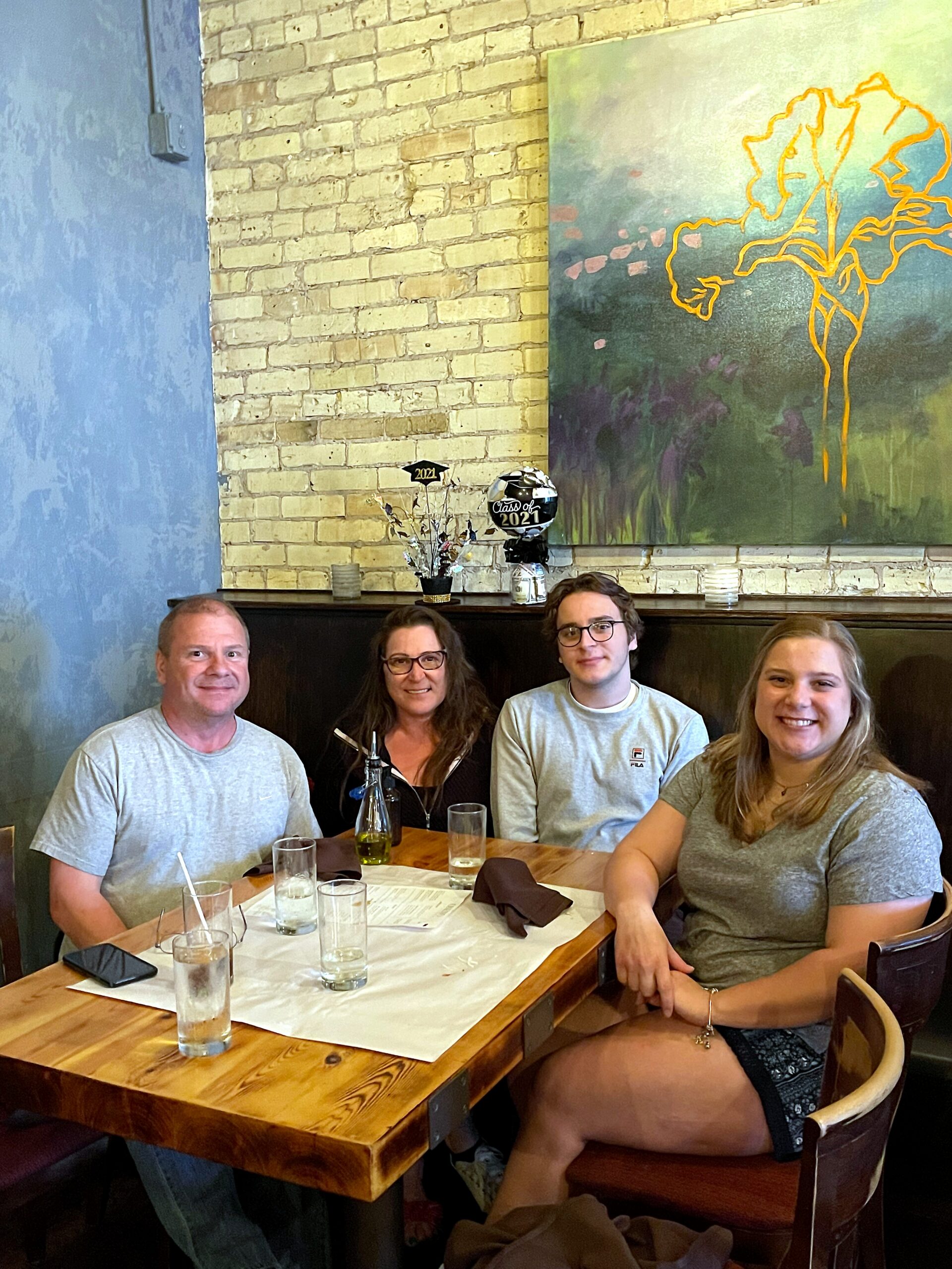 Four people sit around a restaurant table with drinks, posing for a group photo against a brick wall and blue-painted wall art behind them.