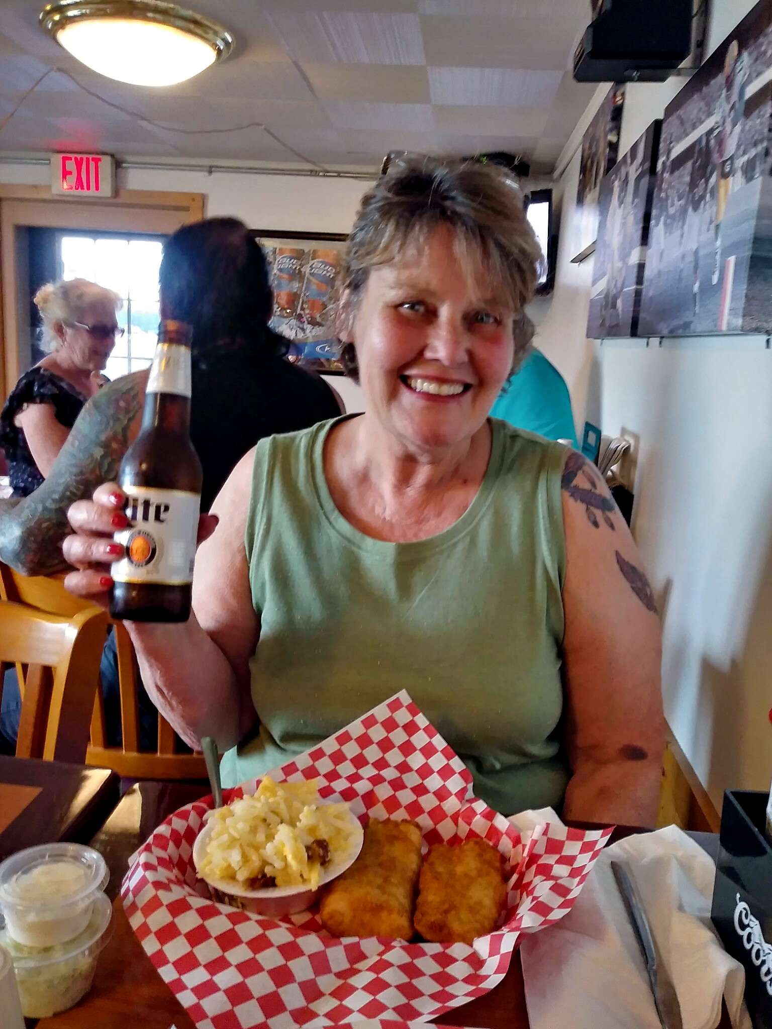 Smiling woman in a green tank top sits at a restaurant table, holding a beer, with a basket of fried foods and mashed potatoes in front.