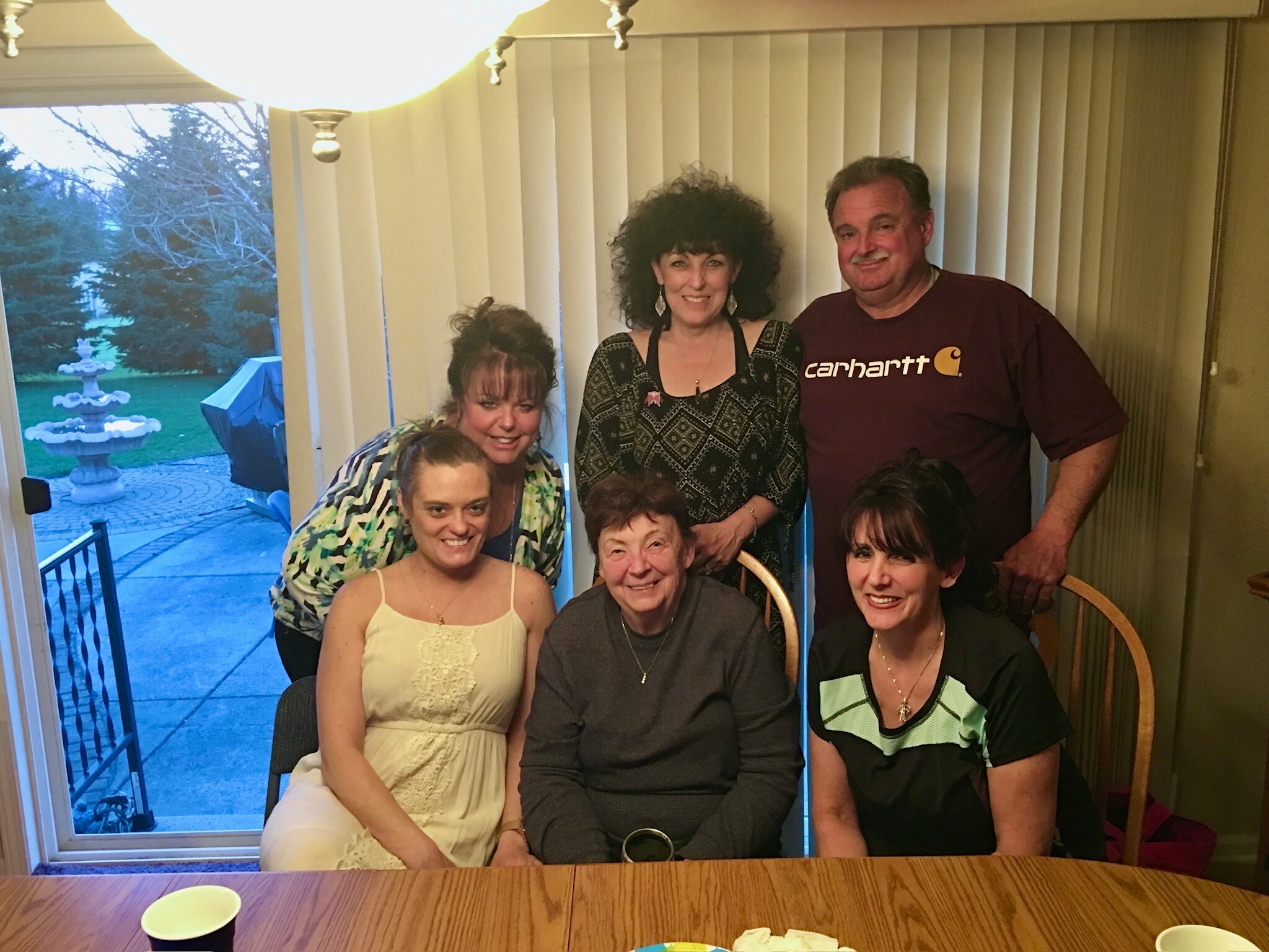 Group of smiling adults gathered around a wooden table inside, posing for a photo with a window and vertical blinds in the background. Outside, a fountain and patio are visible.