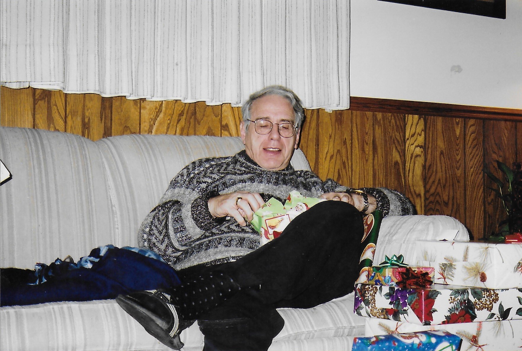 Older man with glasses sits on a striped couch, unwrapping a birthday/holiday gift amid colorful presents in a wood-paneled room.