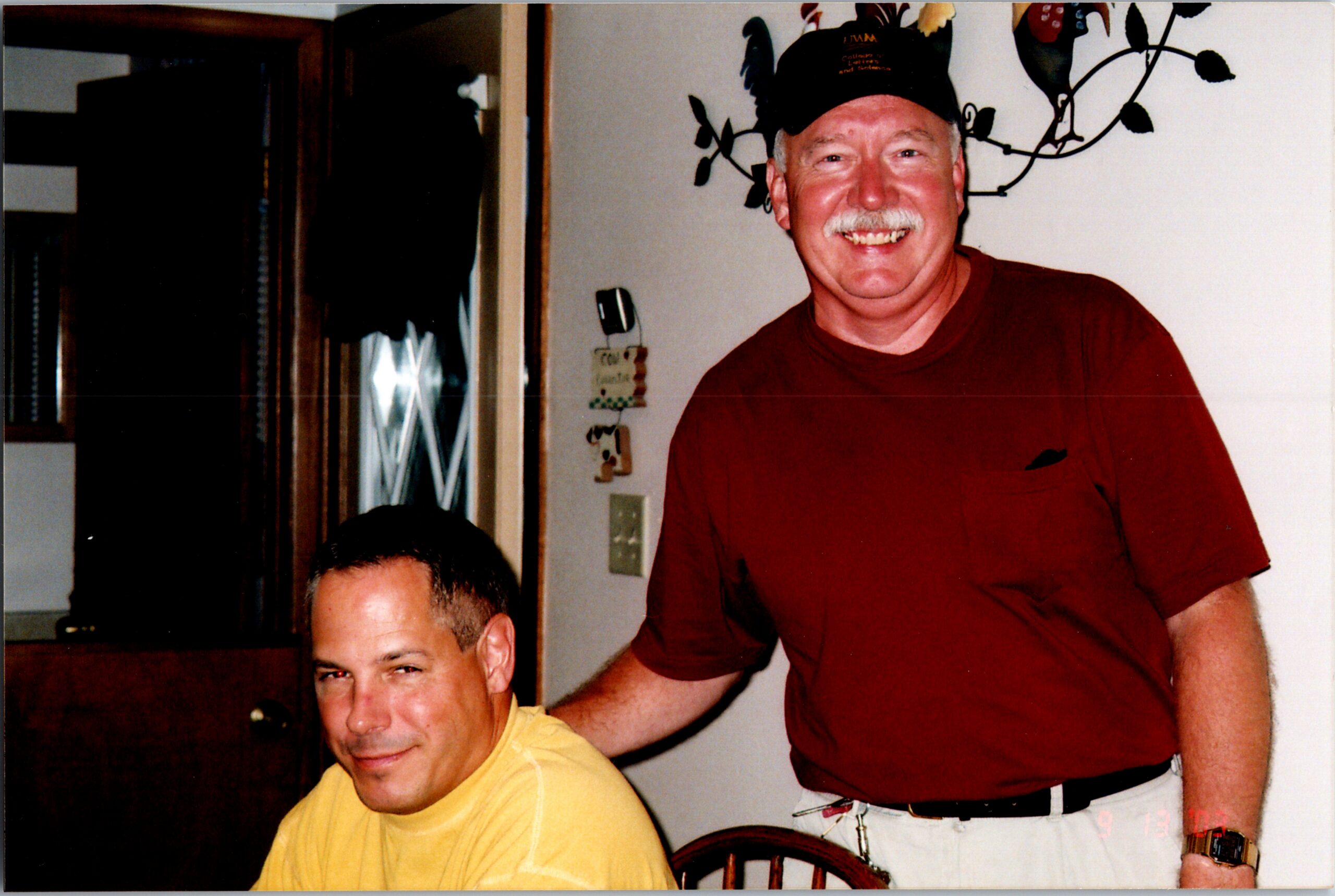 Smiling man in a red shirt and cap stands beside a seated man in a yellow shirt indoors, with wall decor in the background.