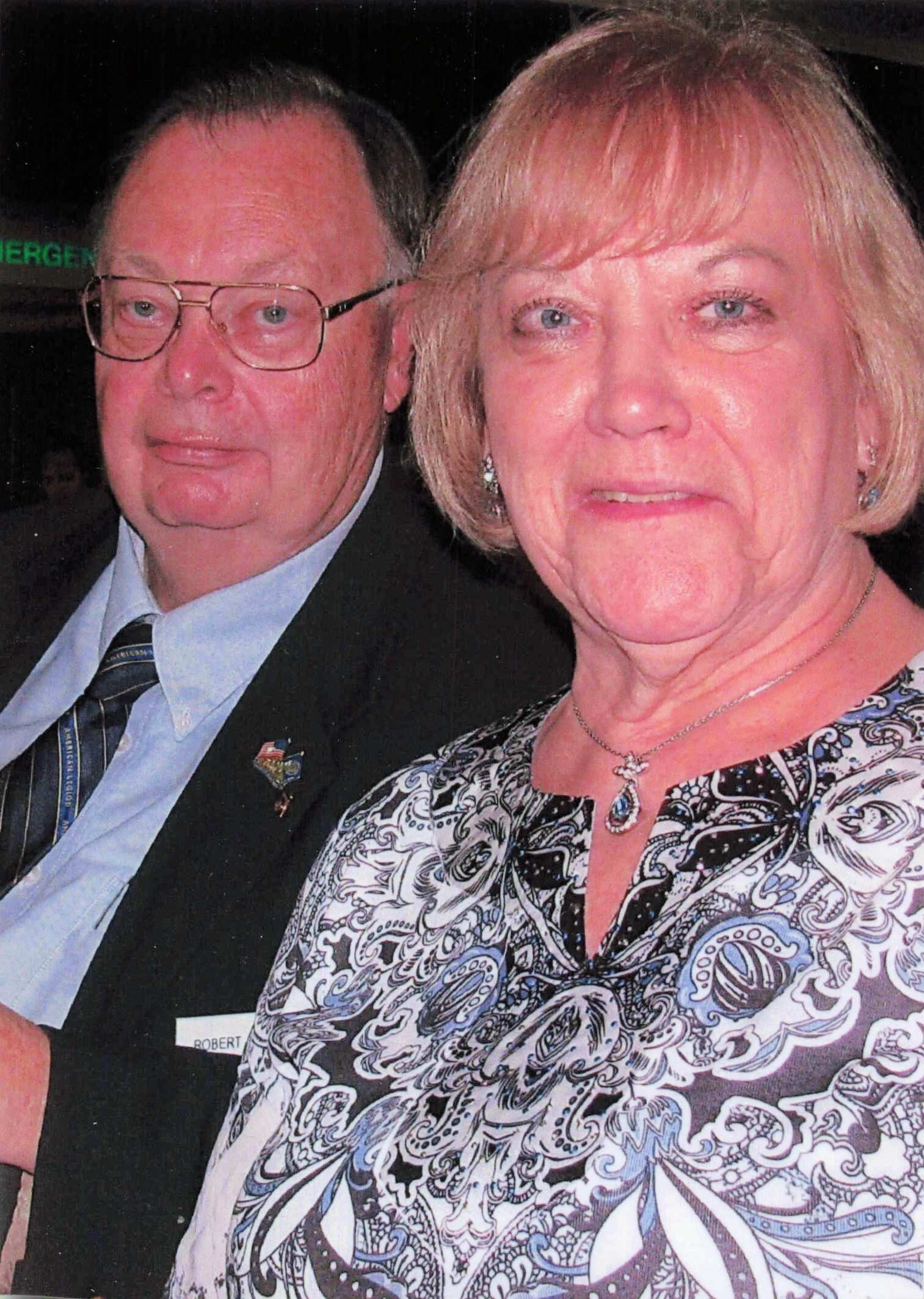 Close-up of an older couple smiling for the camera: man in a suit and glasses beside a woman in a patterned blouse.