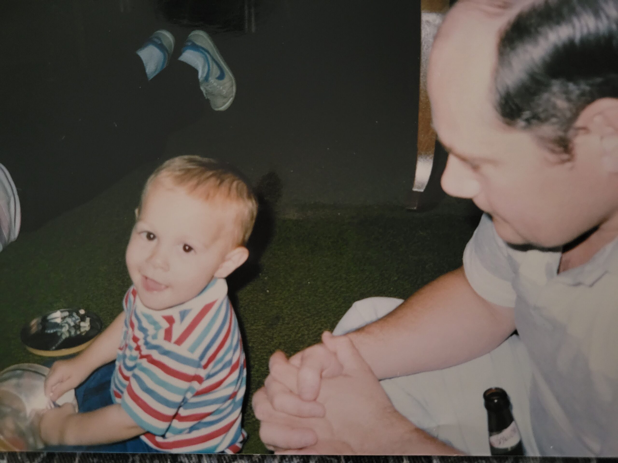 Young child in a red, white, and blue striped shirt sits on grass at night, with an adult beside him and a beer bottle nearby.