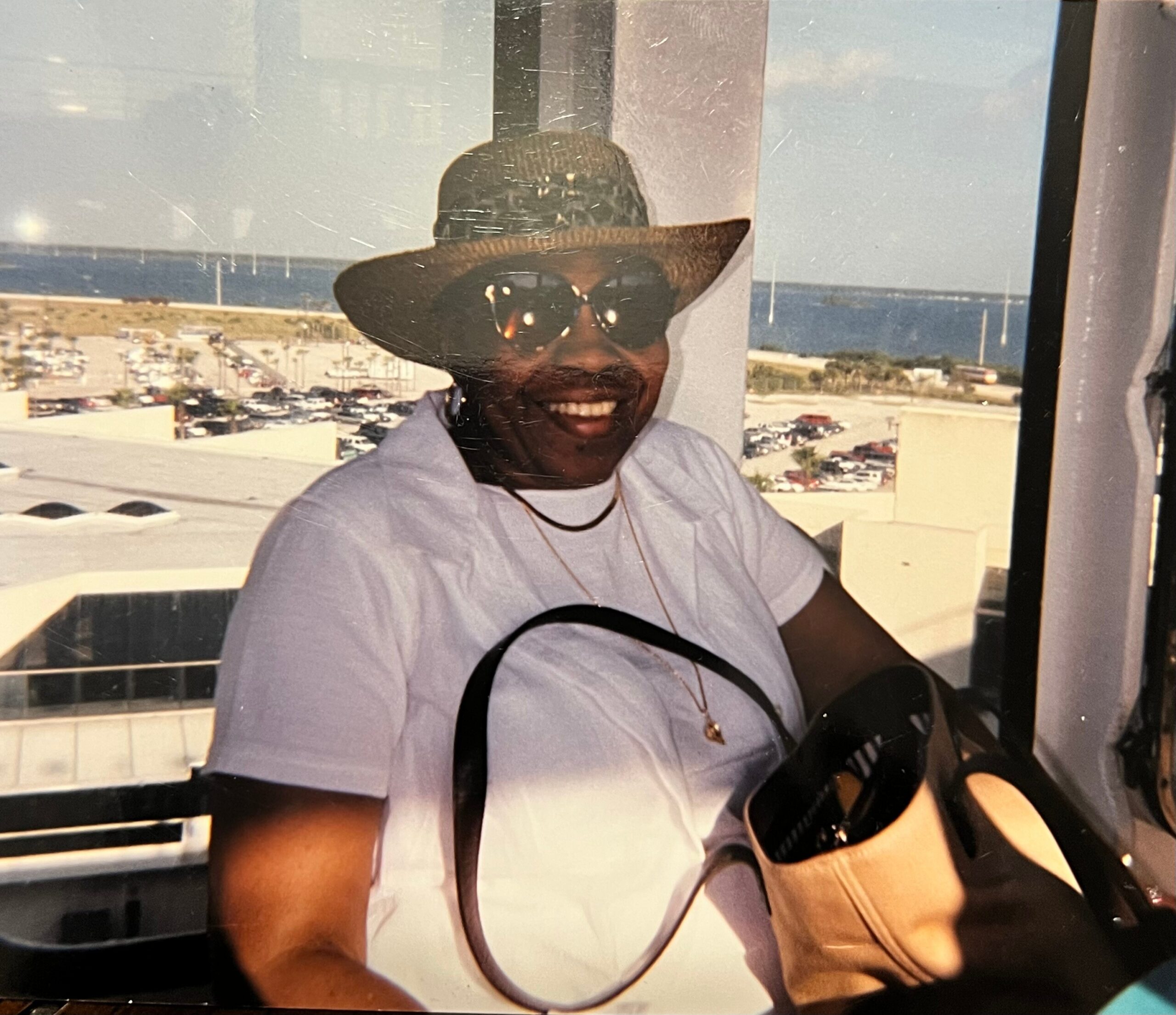 Smiling man wearing a straw hat and sunglasses, playing a guitar, seen as a reflection in a window with a parking lot and coastline in the background