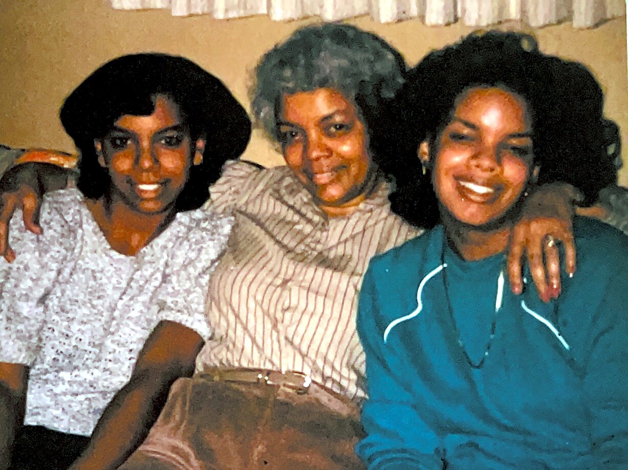 Three smiling women sitting close together, wearing casual outfits, posing for a candid photo.