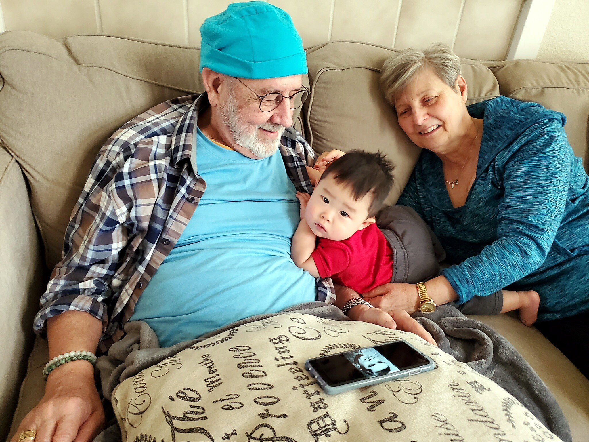Elderly man in a turquoise cap and plaid shirt sits on a beige couch, holding a baby, with a smiling woman in a teal top beside him on the sofa.