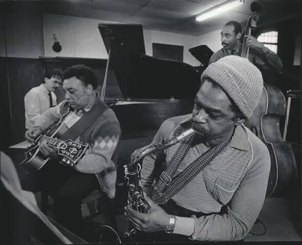 Jazz quartet in a recording studio: saxophonist in a knit cap playing his sax, guitarist beside him, pianist at a grand piano, and a bassist in back.