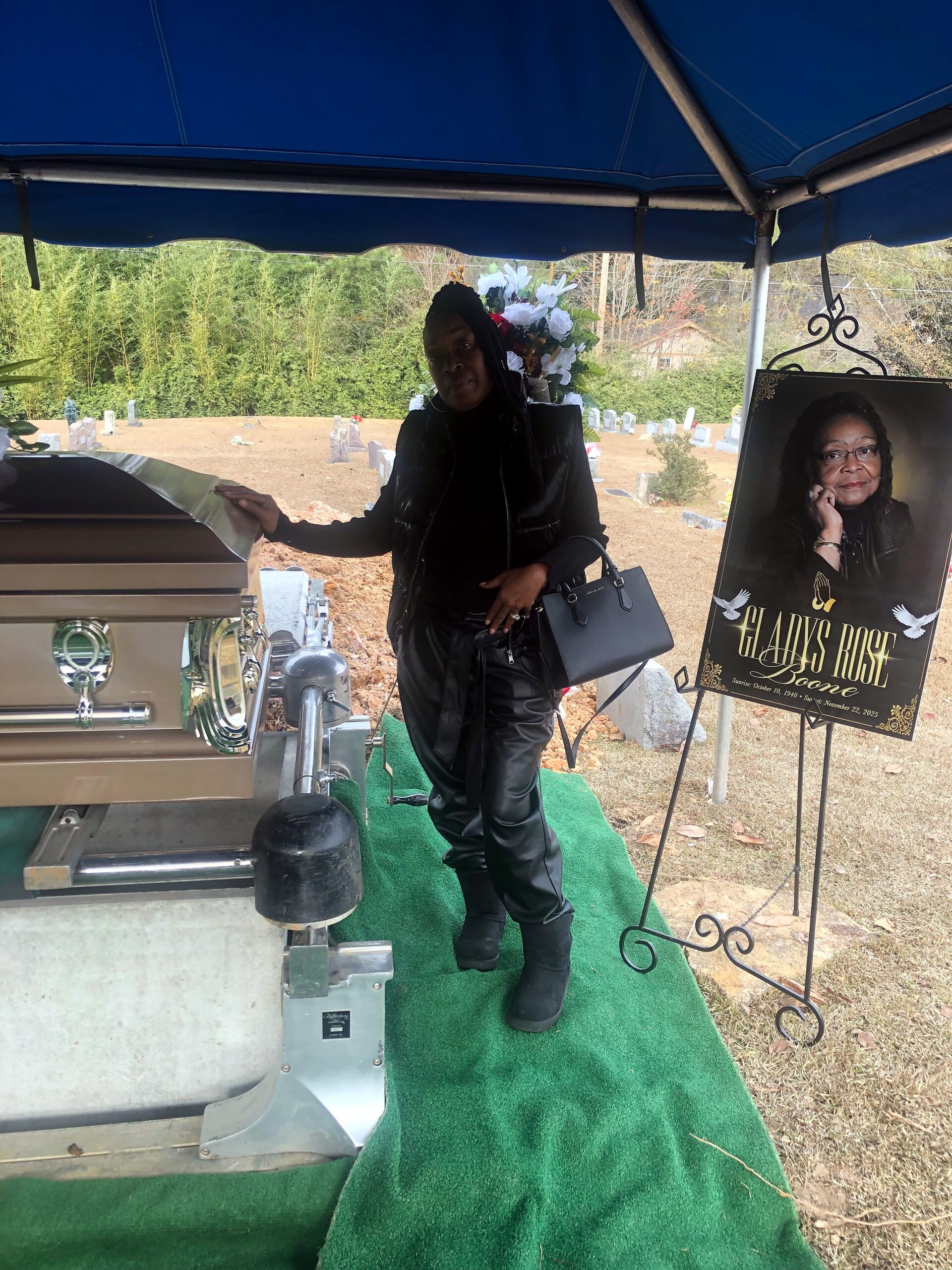 Casket on a wheeled bier under a blue canopy with a woman standing beside, holding a handbag outside a cemetery.