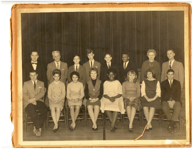 Group of students and teachers posed for a vintage class photo, two rows on a stage with a dark curtain backdrop (sepia-toned).