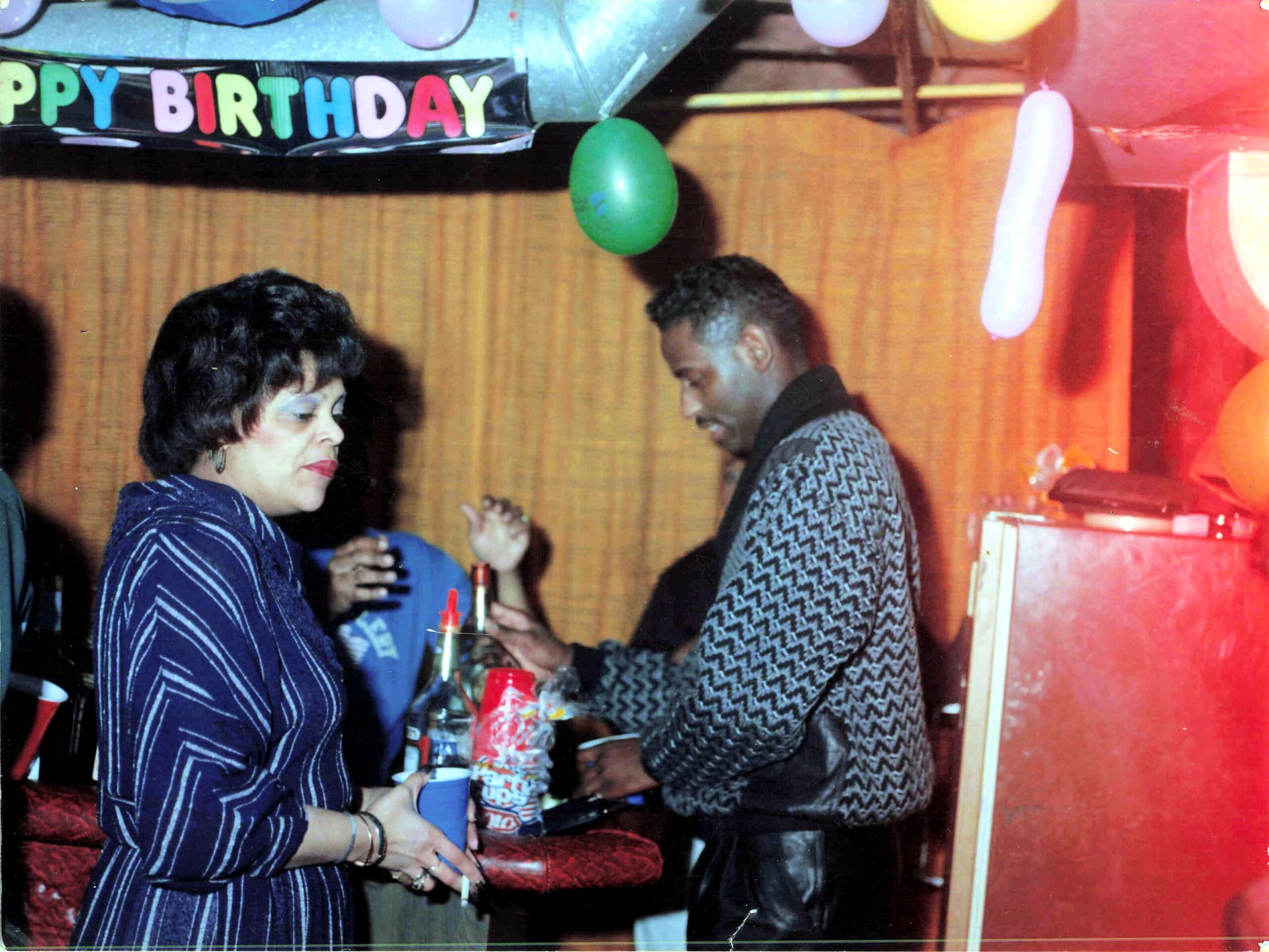 Two people at a birthday party share a toast, with a colorful 'Happy Birthday' banner in the background and drinks on the table.