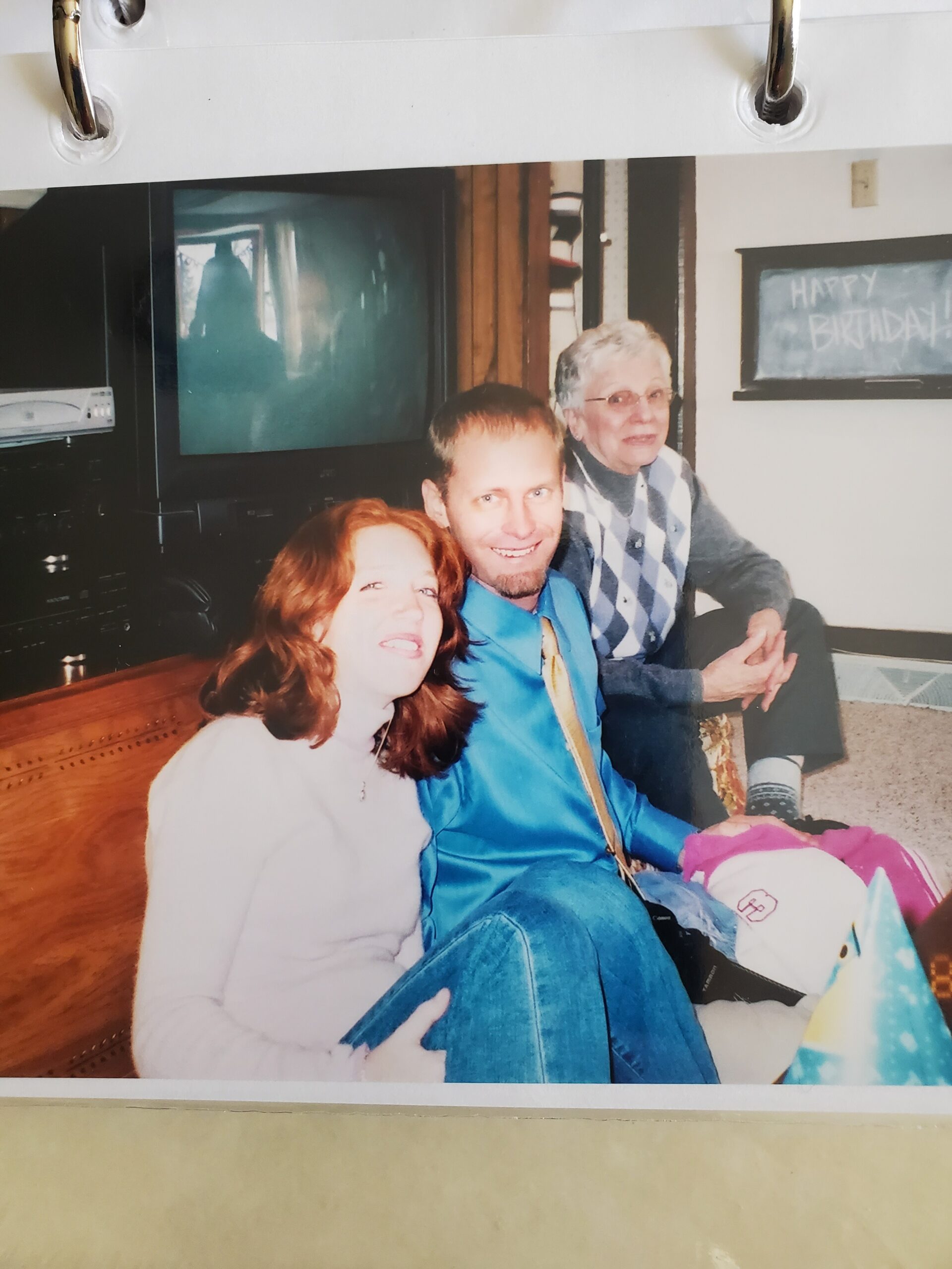 Three people sit close together indoors, smiling at the camera during a birthday gathering, with a chalkboard that says 'Happy Birthday' in the background.
