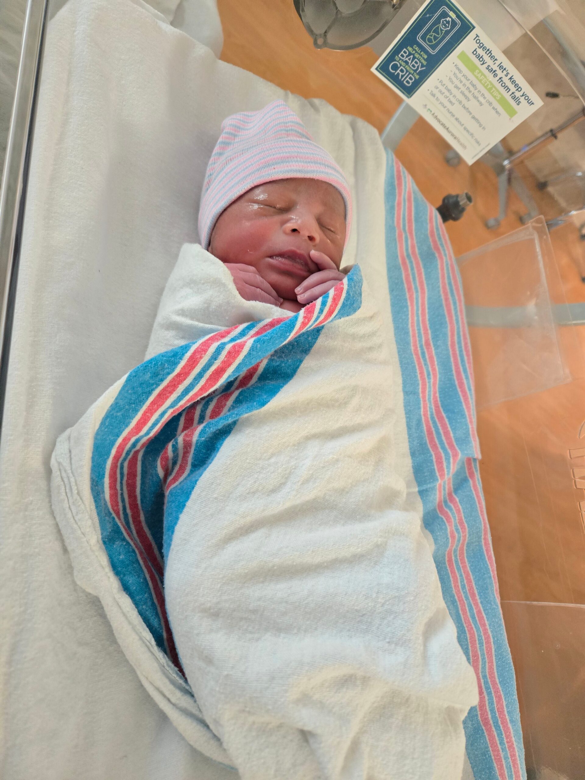 Newborn baby swaddled in a striped blanket wearing a pink-and-blue cap in a hospital incubator.