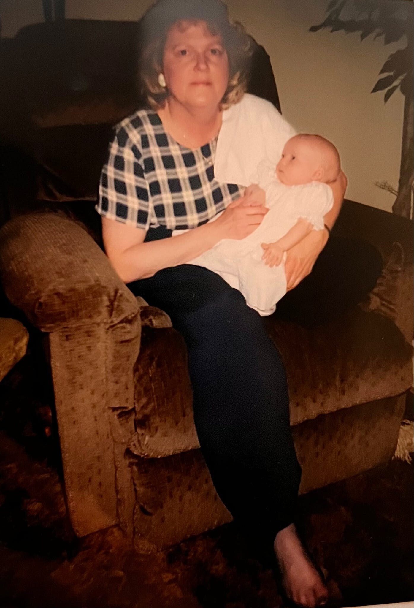 Older woman in a plaid blouse sits on a brown couch, cradling a smiling baby in white clothing on her lap.