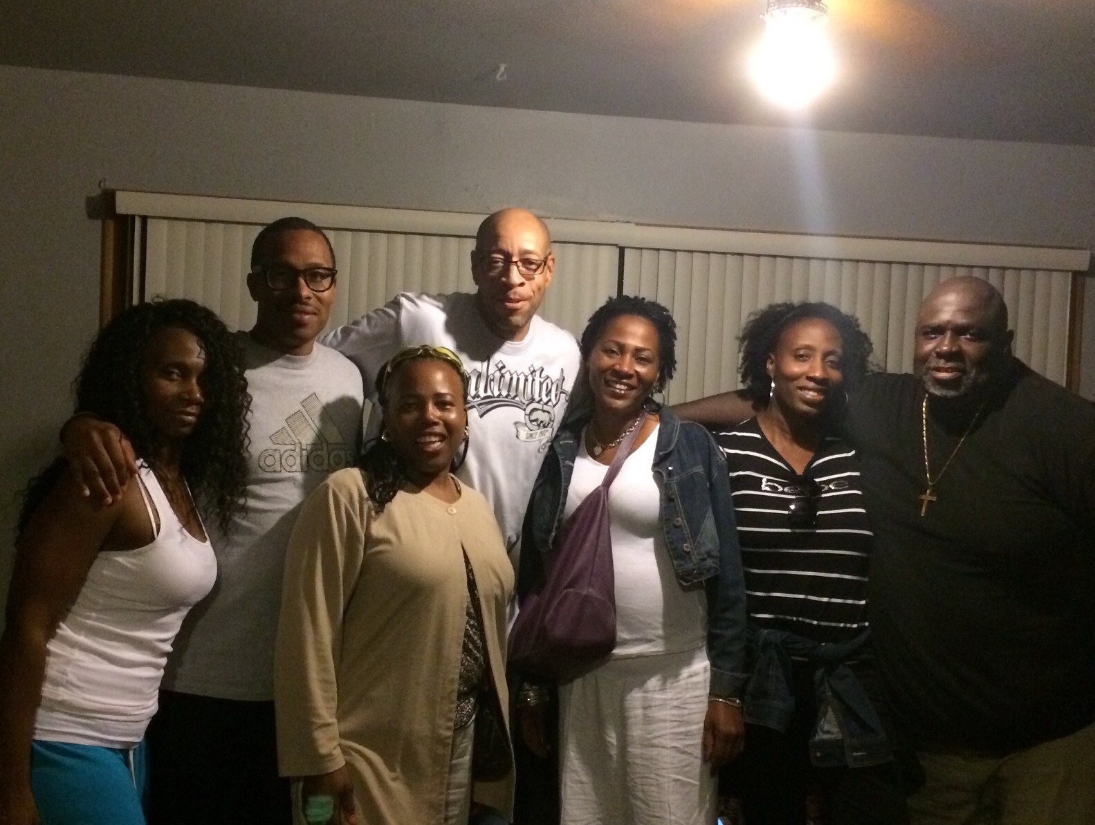Seven adults posing together indoors, smiling for the photo. behind them are vertical blinds on a window.