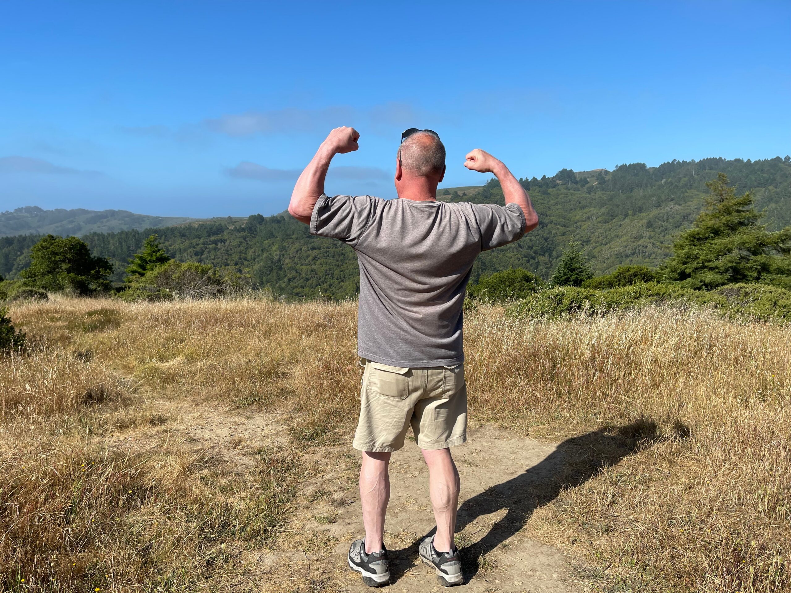 Man standing in a dry field with hills in the distance, arms raised in a flexing pose against a blue sky.