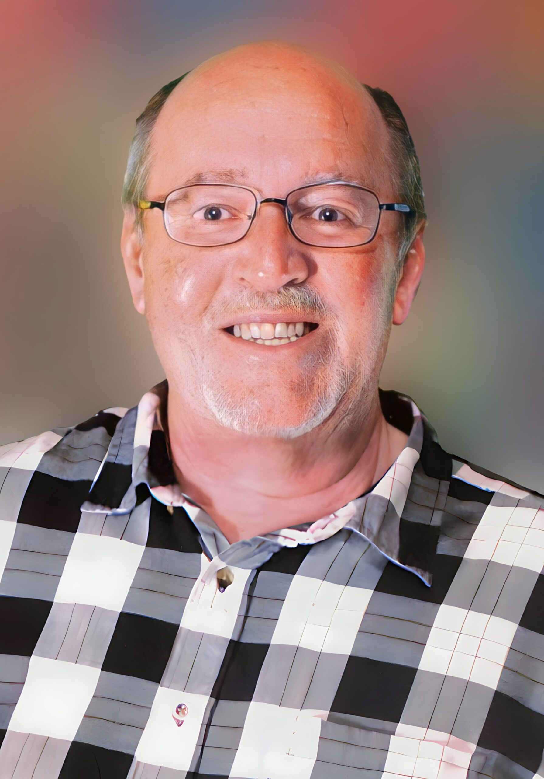 Close-up portrait of a smiling man with glasses, wearing a black-and-white plaid shirt, against a colorful gradient background.