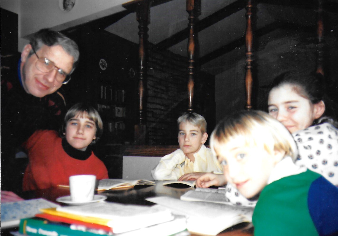 A man with glasses sits beside four children around a table with books, looking toward the camera in a cozy room.
