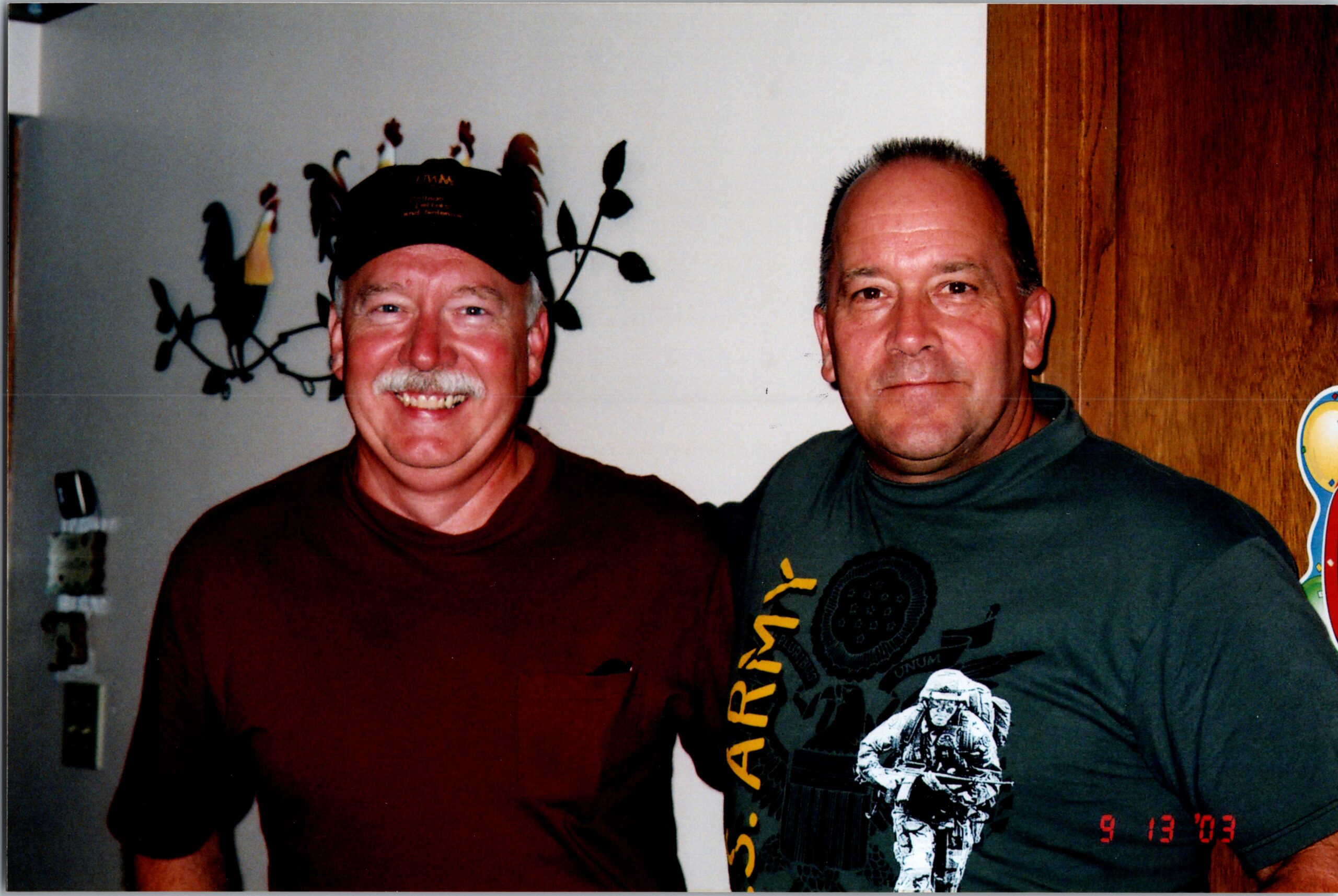 Two men indoors, smiling for a casual photo: left man in a maroon shirt and cap, right man in a gray graphic tee; date stamp 9/13/03 behind them.